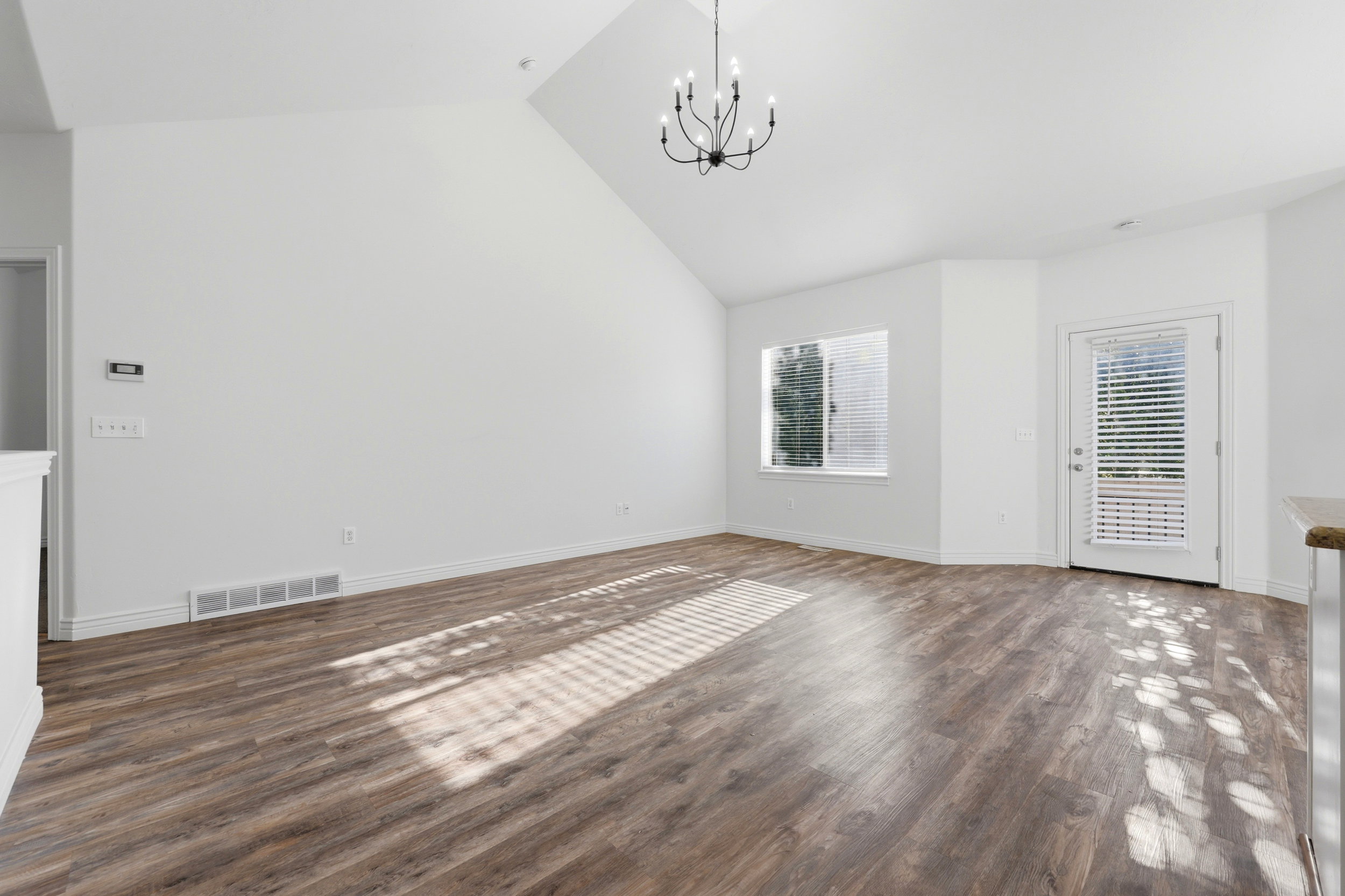 Spacious room featuring vaulted ceilings, wood-finish flooring, a contemporary chandelier, a large window with blinds, and a glass-paneled door