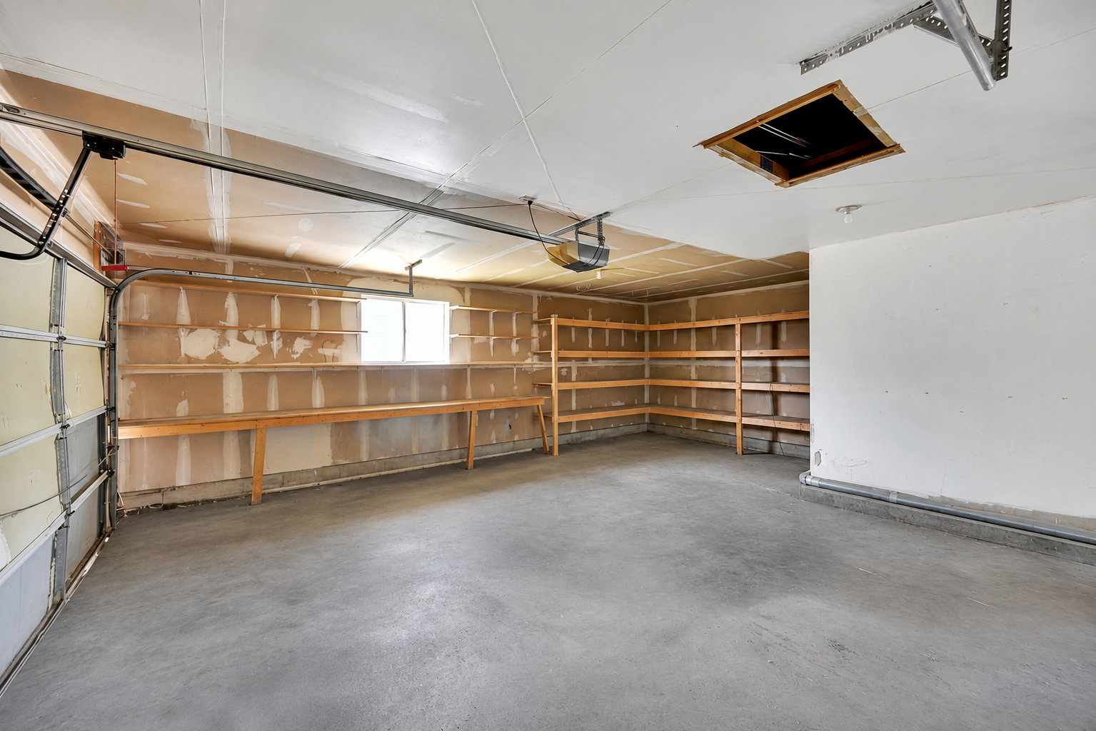 Spacious garage featuring a concrete floor, built-in wooden shelving, a large window, a garage door opener system, and an attic access panel