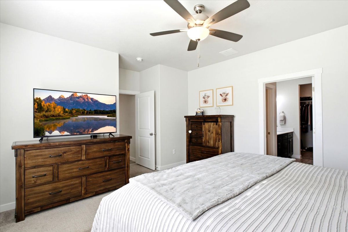 Bedroom featuring neutral wall paint, carpet flooring, a ceiling fan with integrated lighting, and an open doorway revealing a bathroom vanity and closet