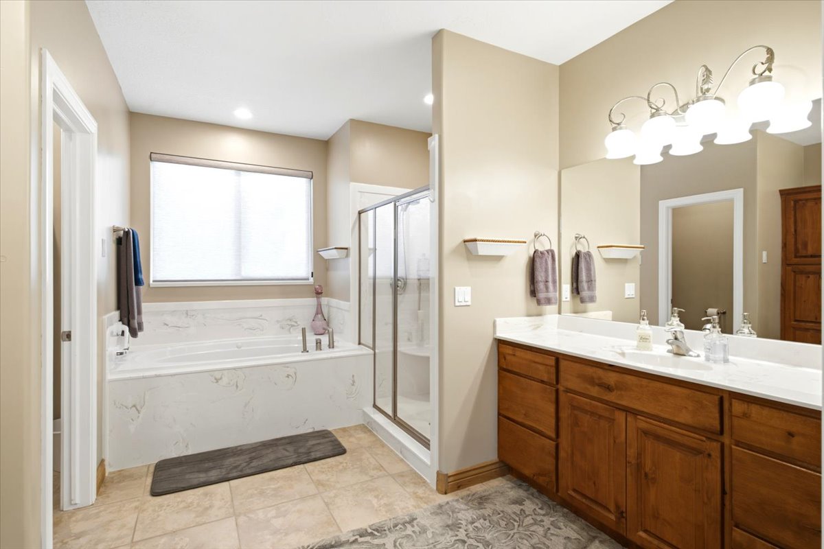 Bathroom featuring a double vanity with wood cabinetry and light-toned countertops, a built-in soaking tub with a tiled surround, a glass-enclosed shower, tile flooring, and recessed lighting