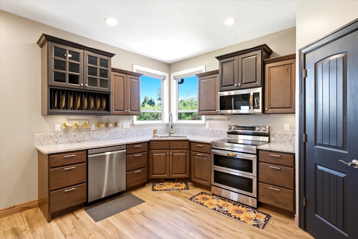 Kitchen featuring wood-finish flooring, two large windows, granite-patterned countertops, wood cabinetry with varied finishes, and stainless steel appliances