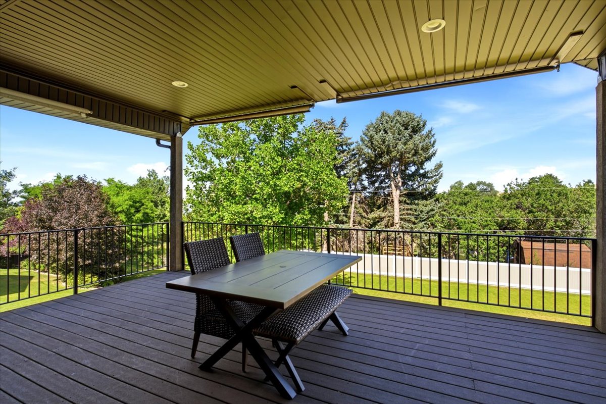 Expansive covered deck featuring composite decking, black metal railings, a paneled ceiling with recessed lighting, and a verdant tree-lined perimeter
