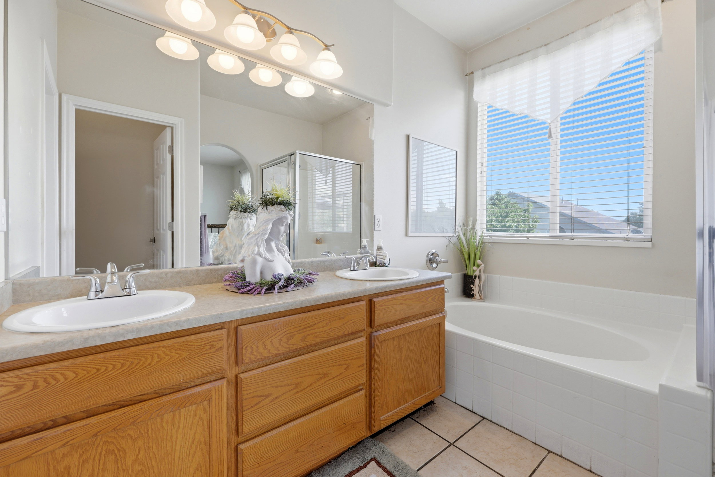 Bathroom featuring a double vanity with wood-finish cabinetry, a drop-in soaking tub, a large mirrored wall, and tiled flooring