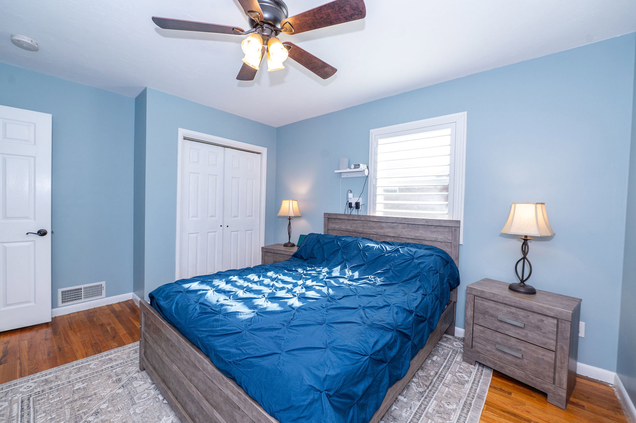 Room with wood-finish flooring, a ceiling fan with integrated lighting, and a window featuring louvered shutters