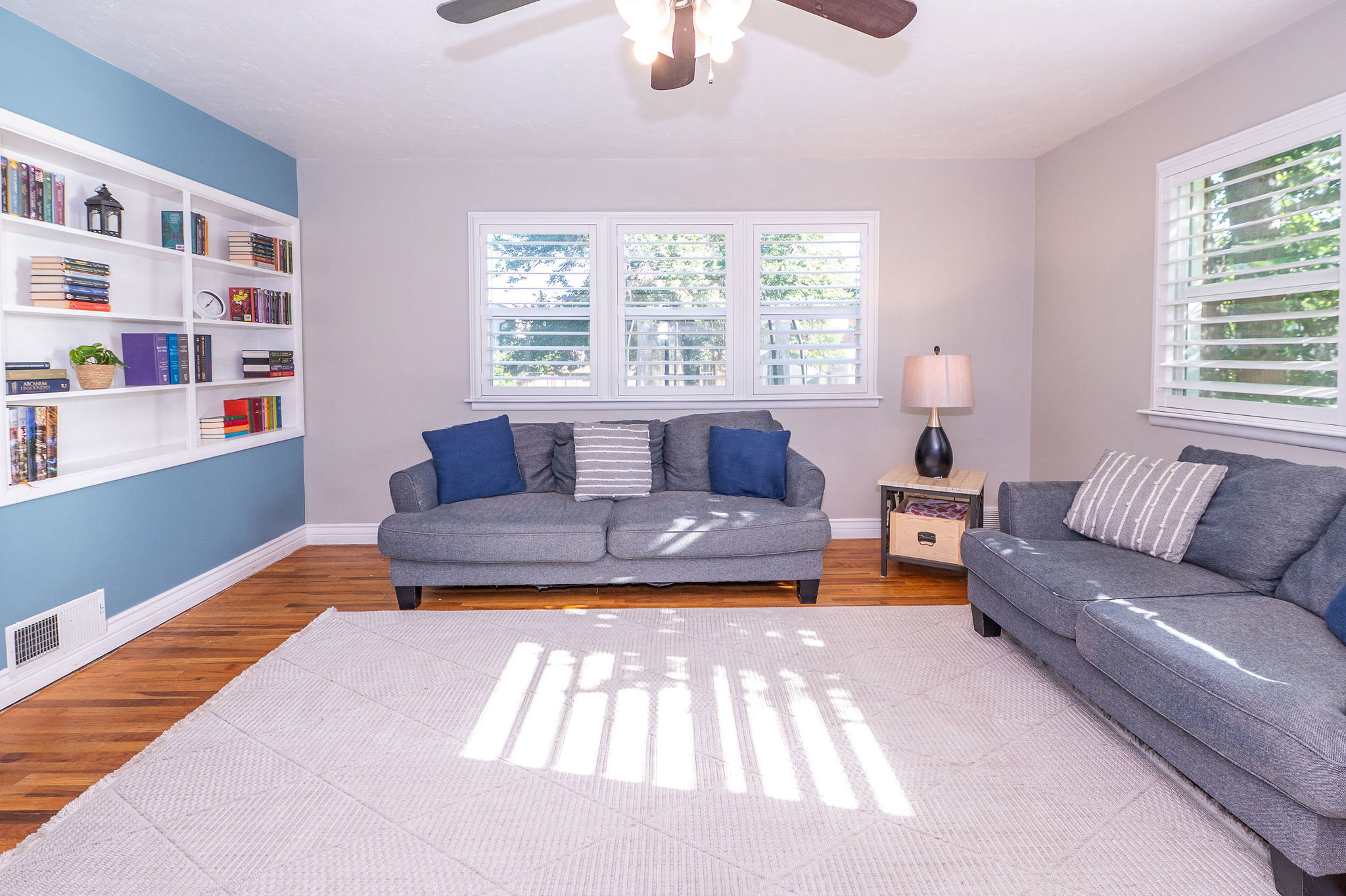 Spacious living area featuring wood-finish flooring, built-in shelving, white plantation shutters, and a ceiling fan
