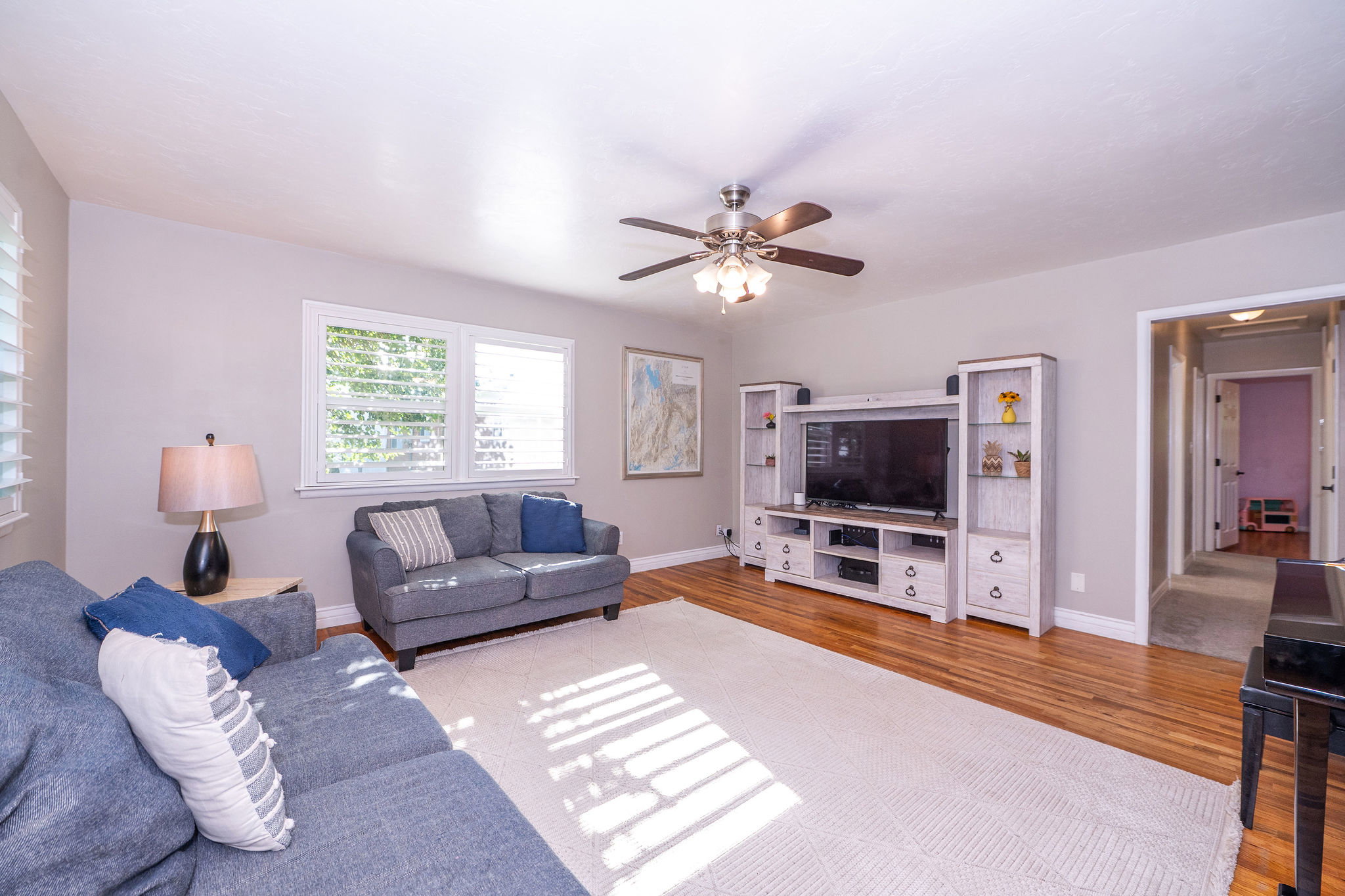 Spacious living area featuring polished wood-finish flooring, multiple windows with white louvered blinds, and a ceiling fan with integrated lighting