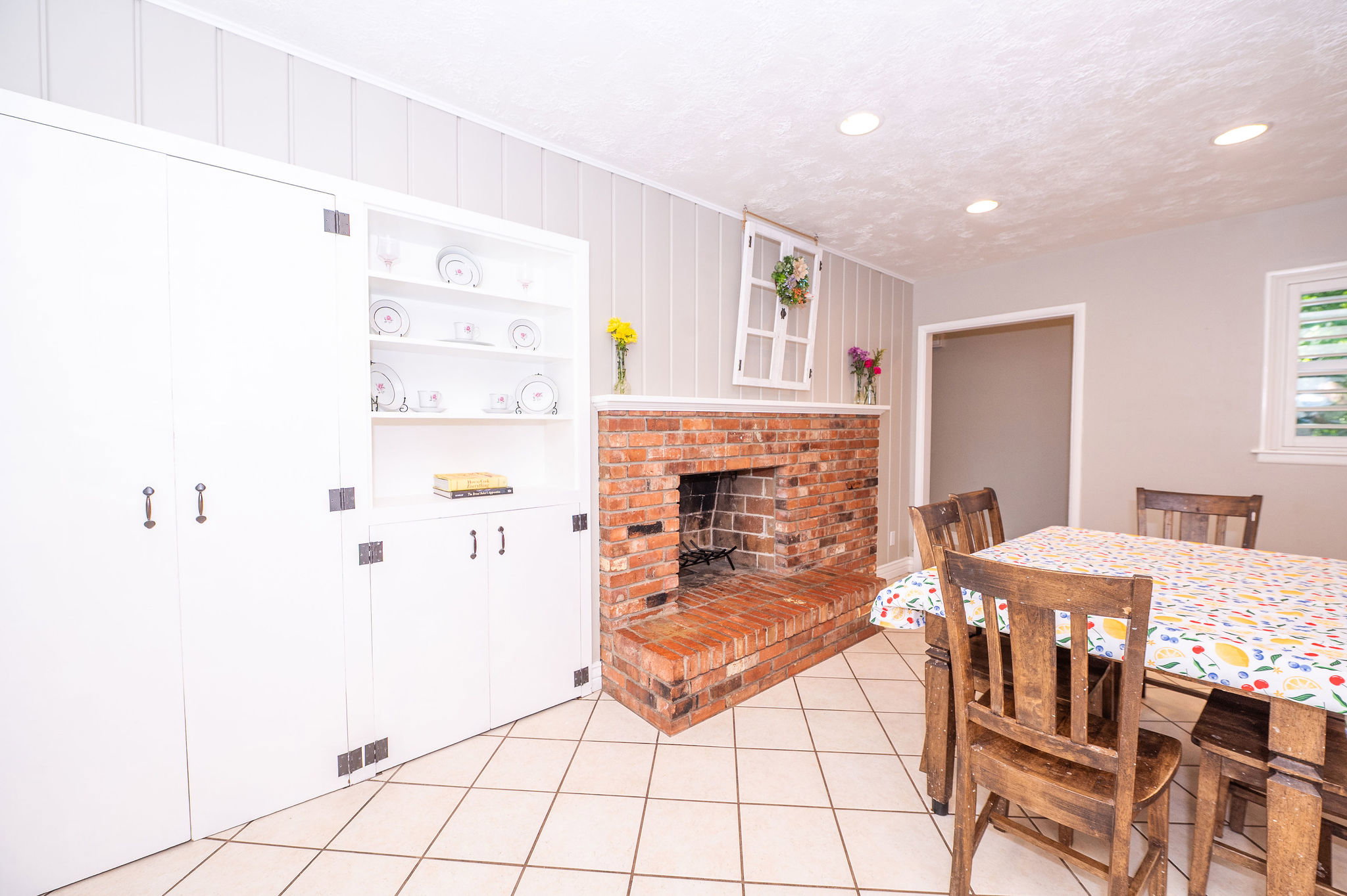Tile flooring, a brick fireplace, white built-in cabinetry with open shelving, recessed lighting, and a window with white louvered shutters