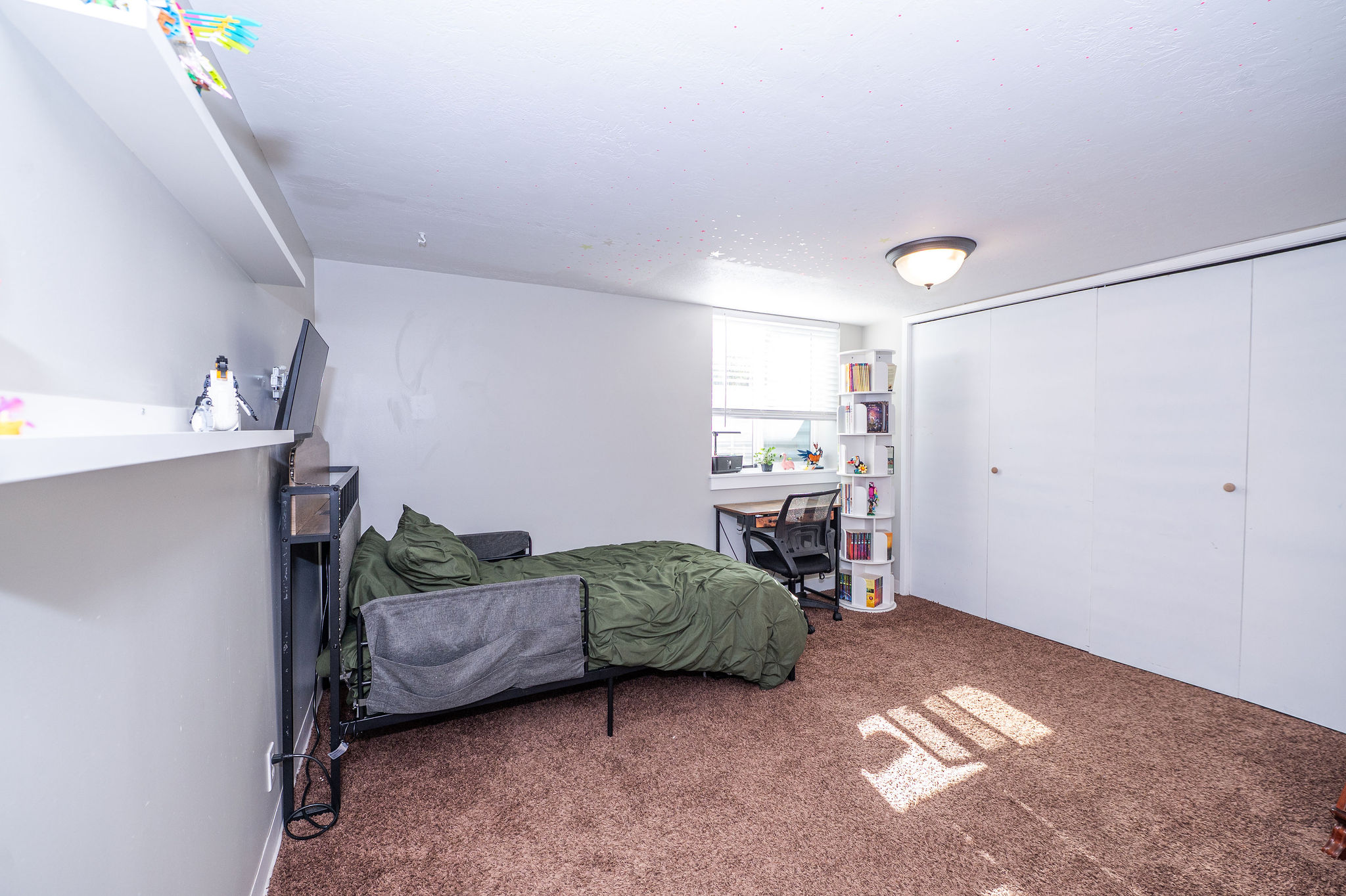 Wall-to-wall carpeting in a rich brown hue, bright window with blinds, overhead flush-mount lighting, and bi-fold closet doors