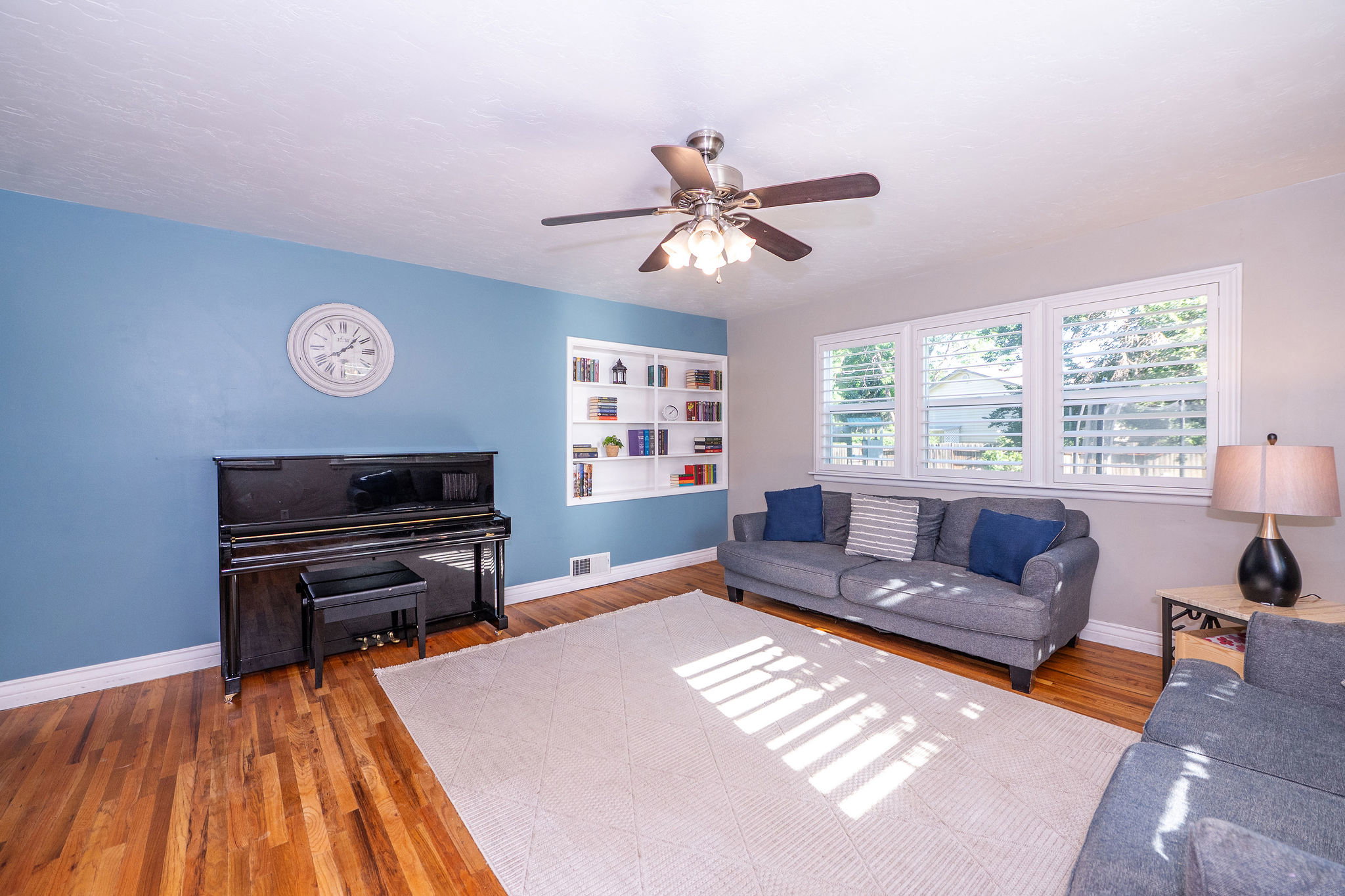 Hardwood flooring complements a room featuring a ceiling fan with integrated lighting, multiple windows with white plantation shutters, and a built-in shelving unit