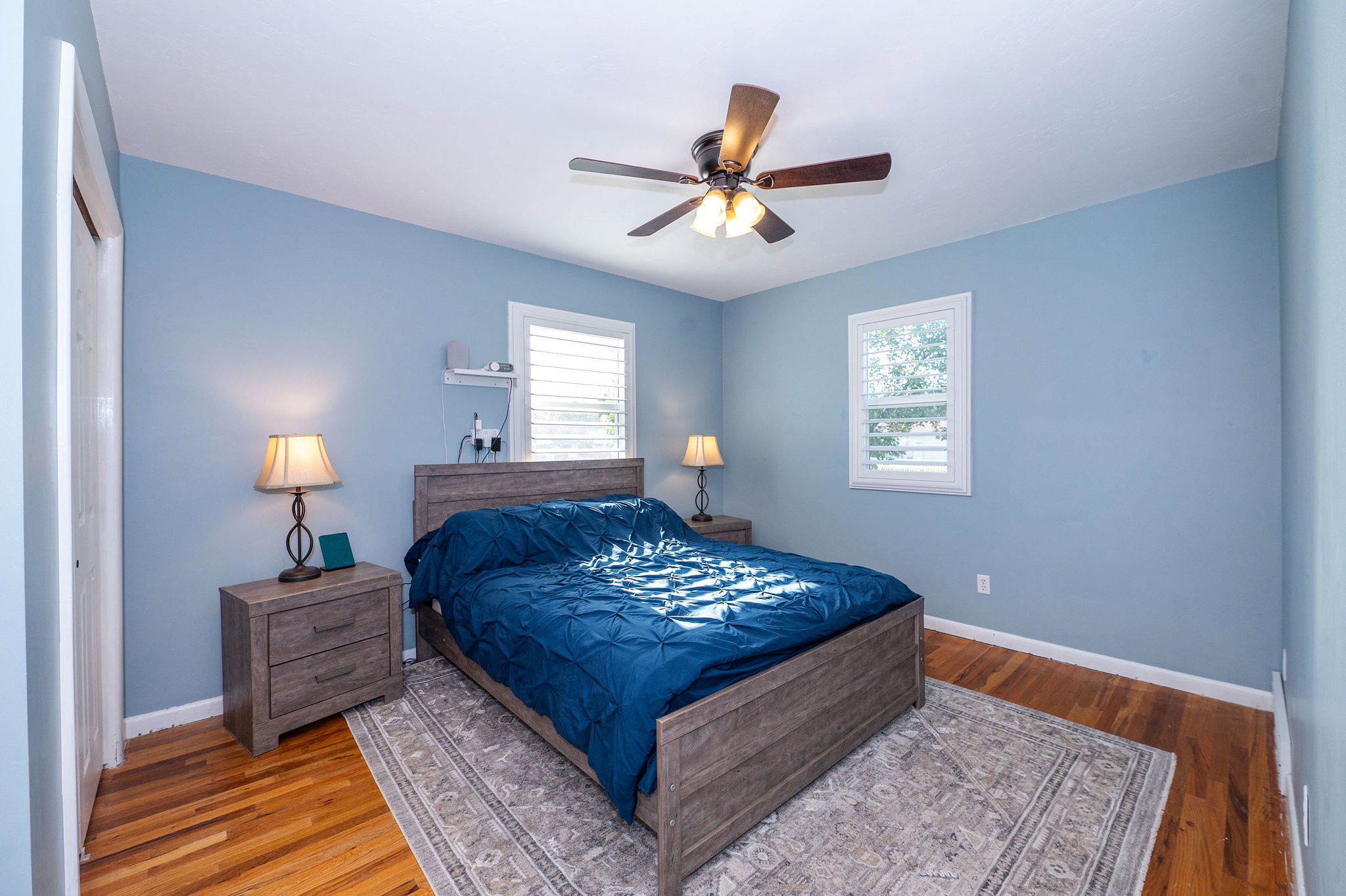 Bedroom featuring wood-finish flooring, two windows with plantation shutters, and a ceiling fan with integrated lighting