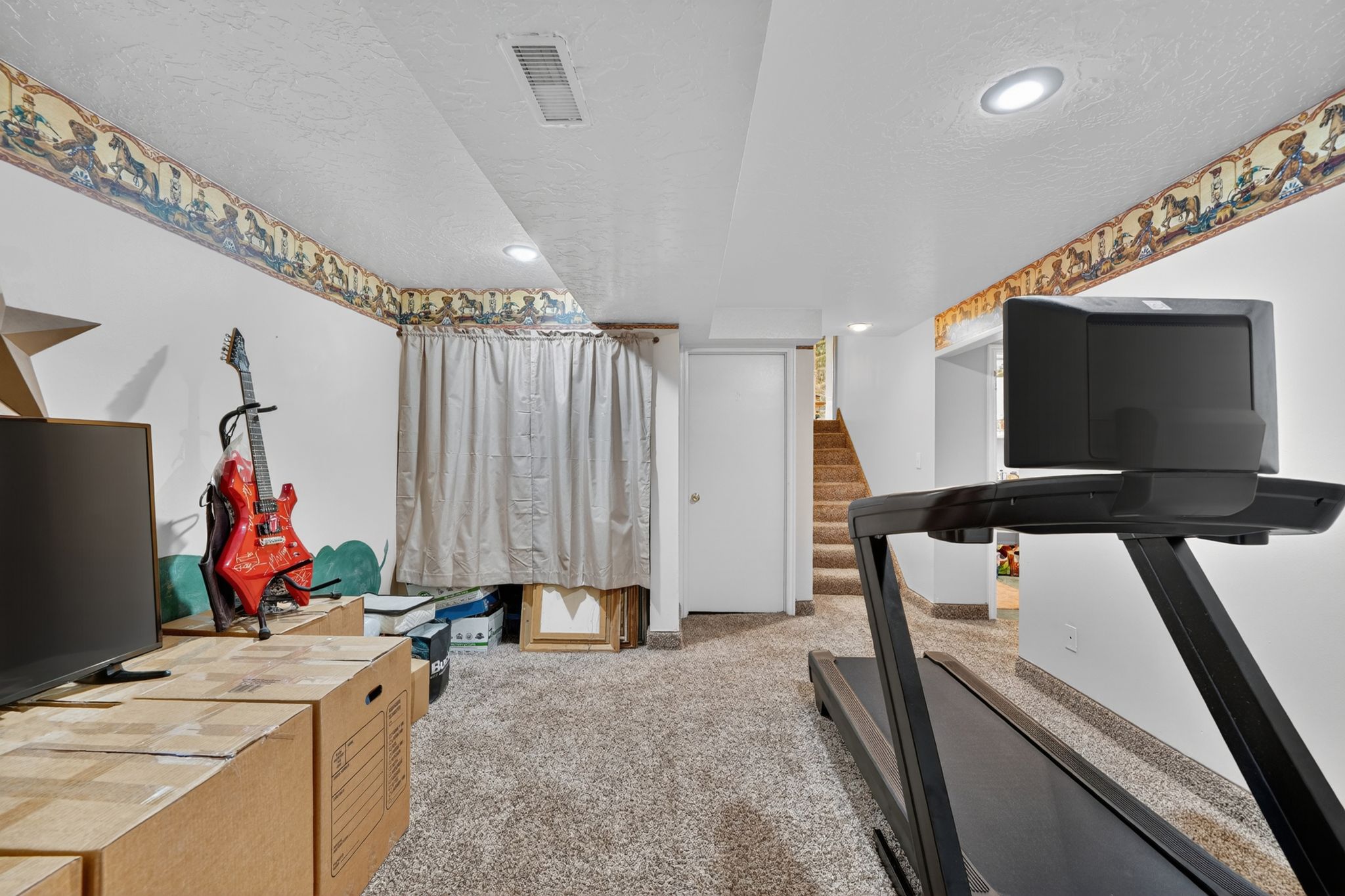 Carpeted lower-level room featuring recessed lighting, white walls with decorative border, a closed white door, and a carpeted staircase