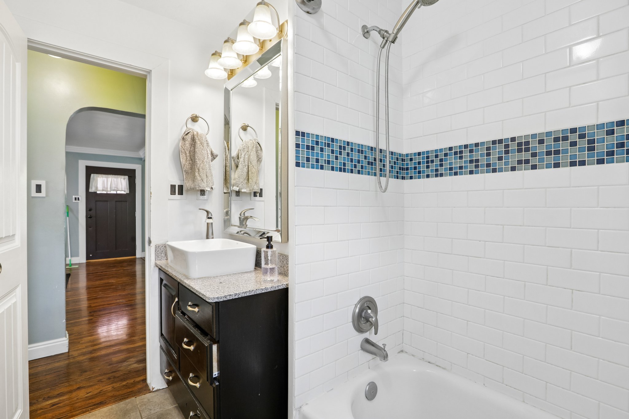 Bathroom featuring a white subway tile shower with a blue mosaic accent band, a white rectangular vessel sink, a dark wood-finish vanity with drawer storage, and a light-colored countertop