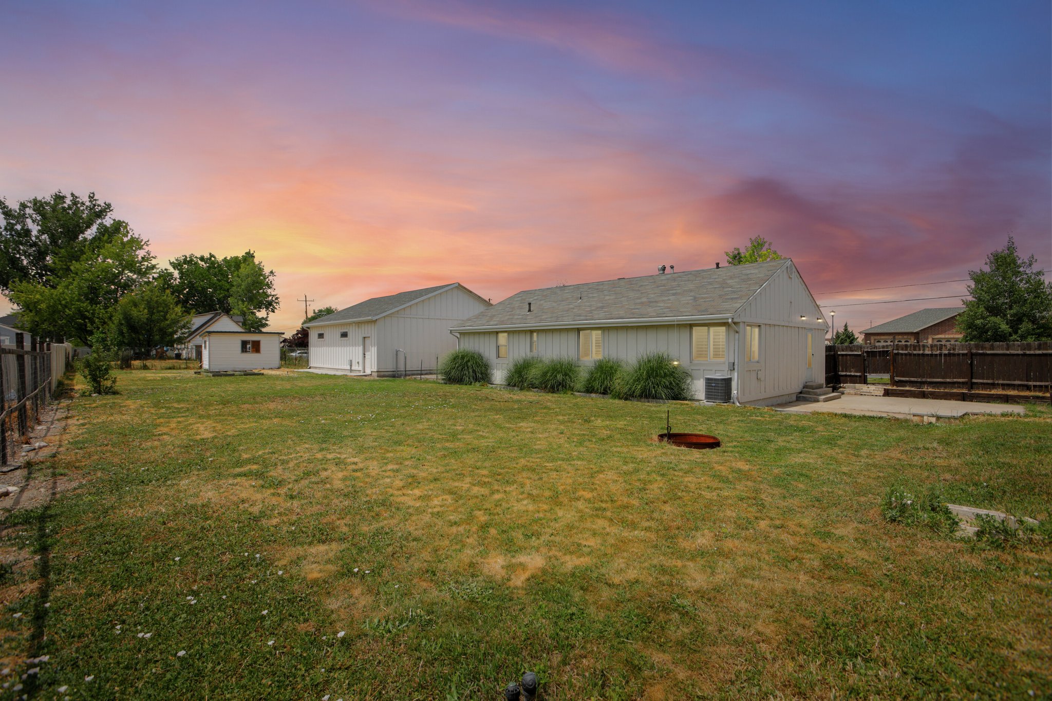 Spacious backyard featuring a single-story residence with light-colored siding, a detached garage, a separate shed, and a concrete patio area