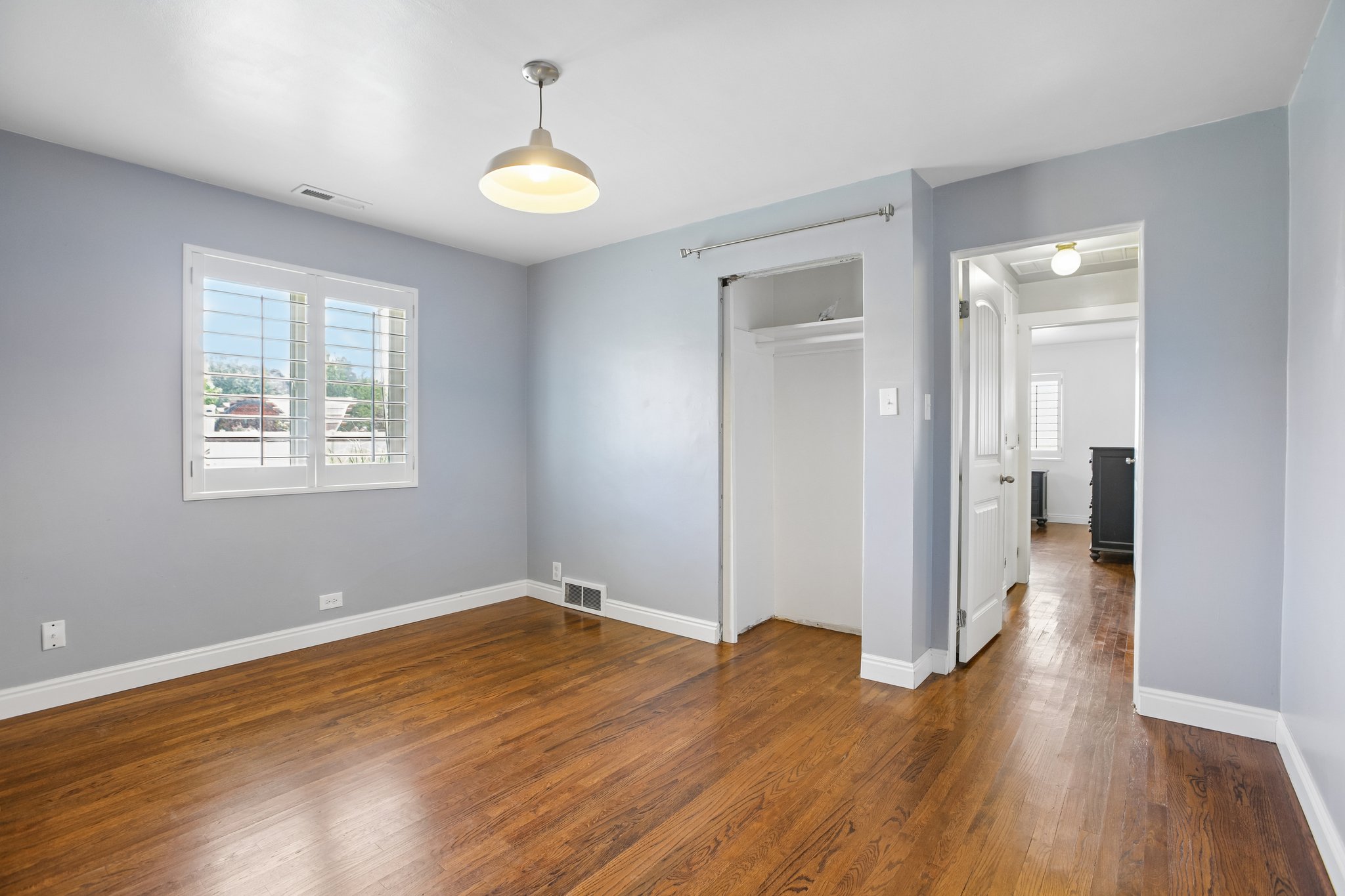 Room featuring wood-finish flooring, a window with white louvered shutters, a built-in closet with hanging rod, and a contemporary pendant light fixture