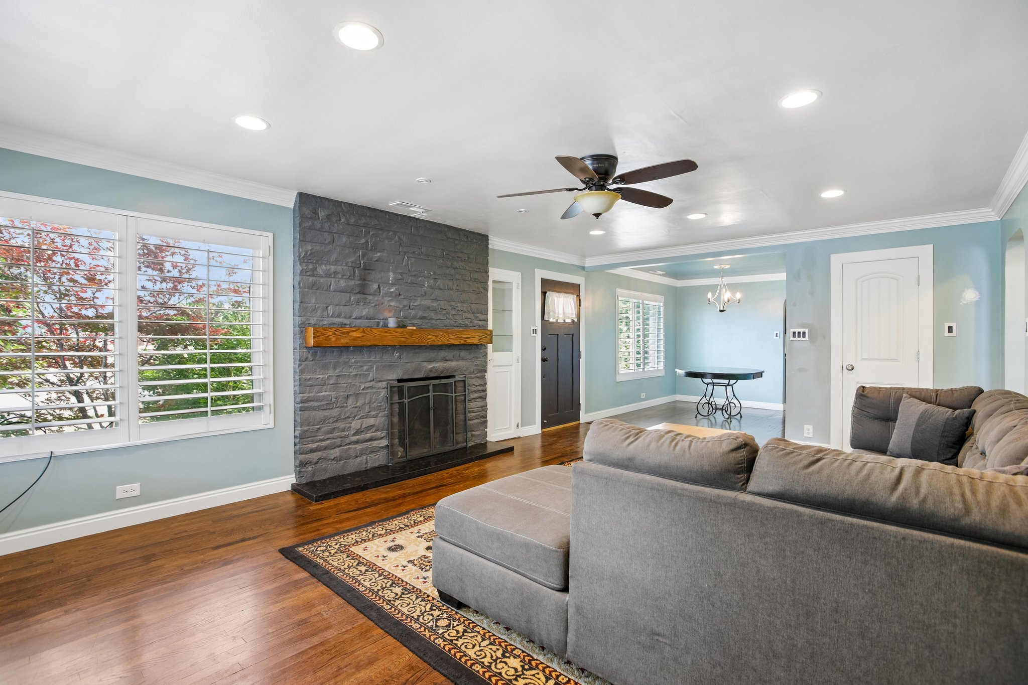 Living area featuring wood-finish flooring, a stone-clad fireplace with a wood mantel, and multiple windows with plantation shutters