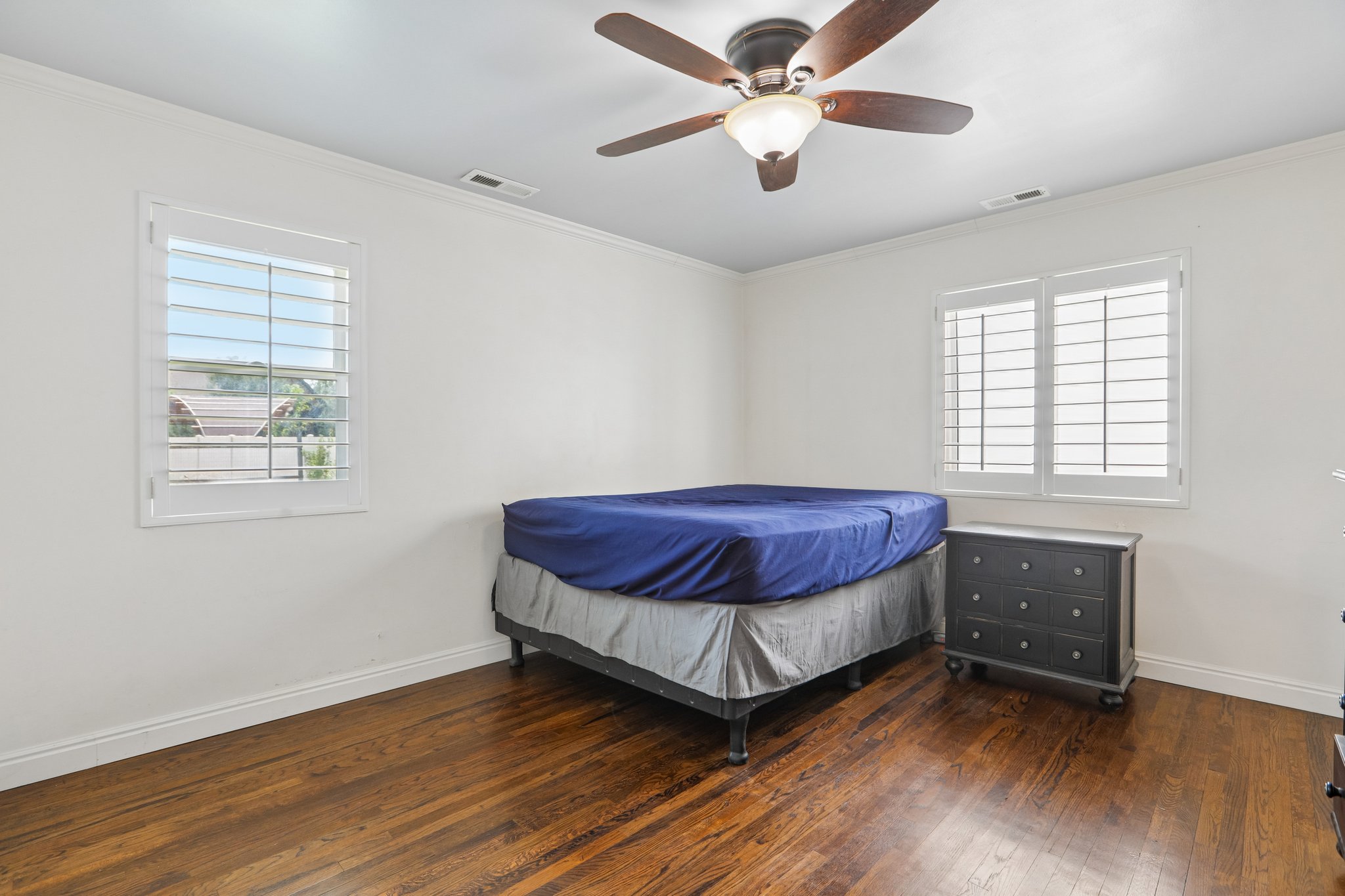 Bright room featuring hardwood flooring, white walls, two windows with white plantation shutters, and a ceiling fan with integrated lighting