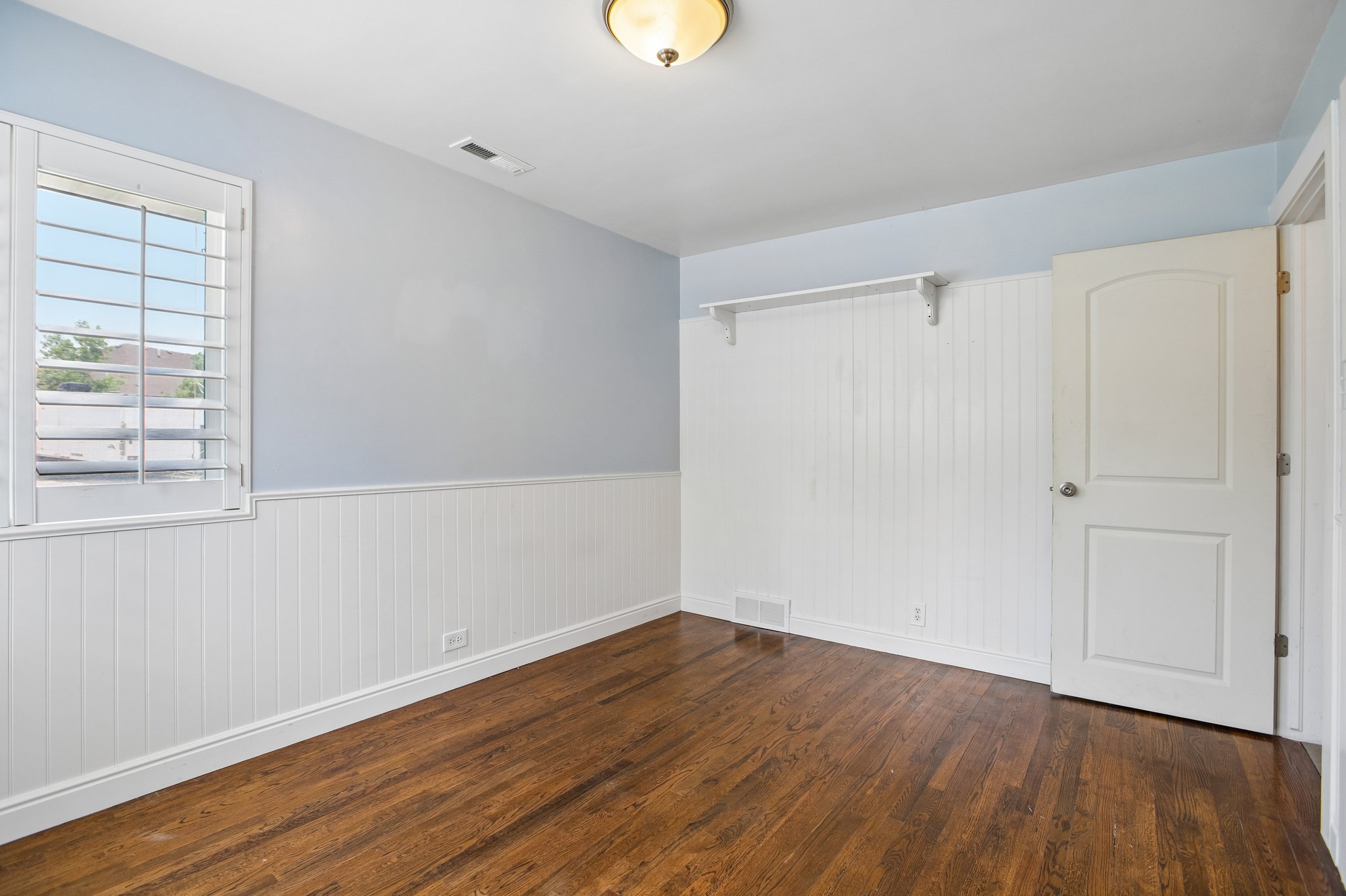 Room featuring wood-finish flooring, wainscoting, and a window with white louvered shutters