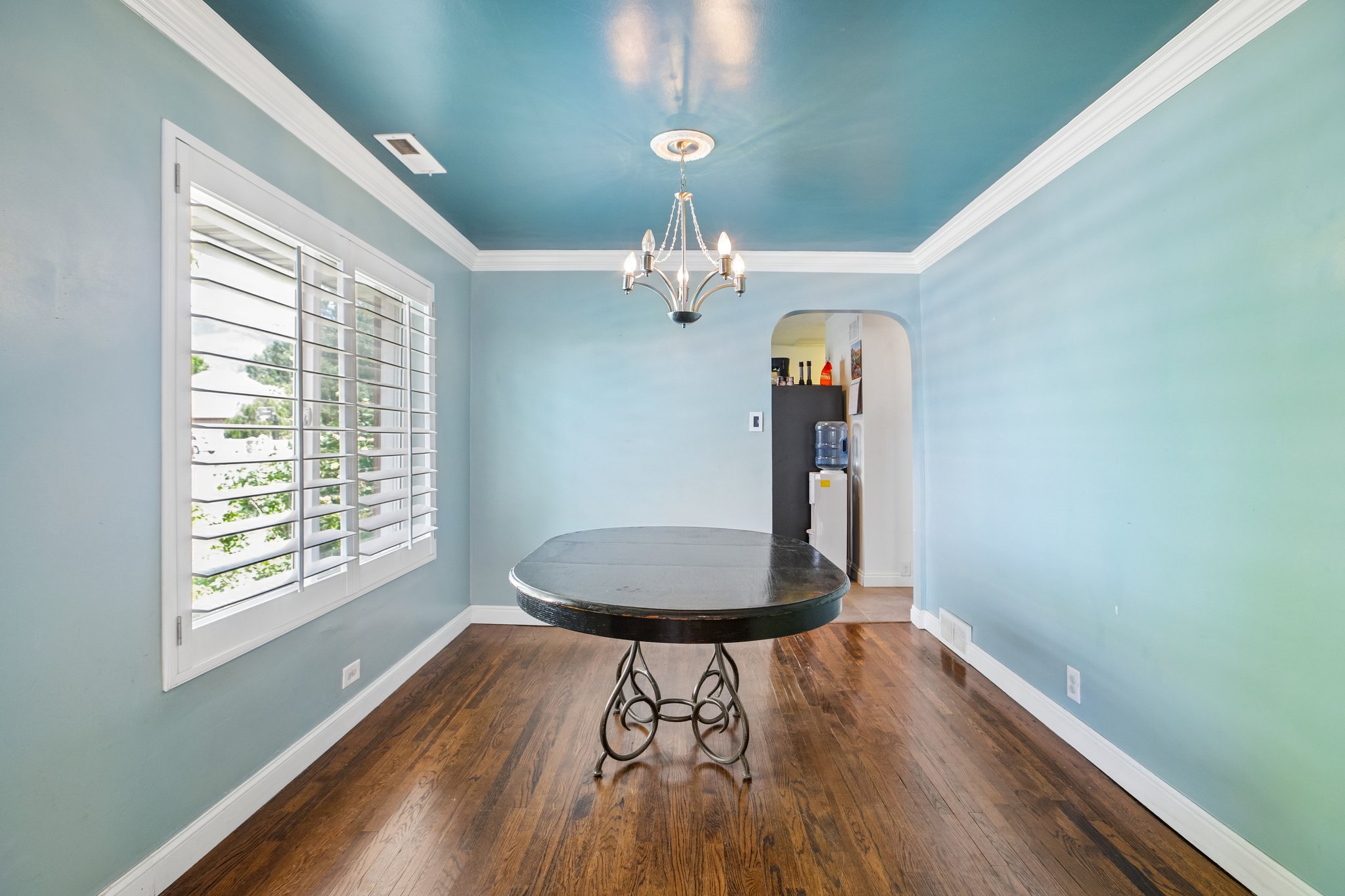 Bright room featuring rich wood-finish flooring, white crown molding, a decorative chandelier, and a wide window with white plantation shutters