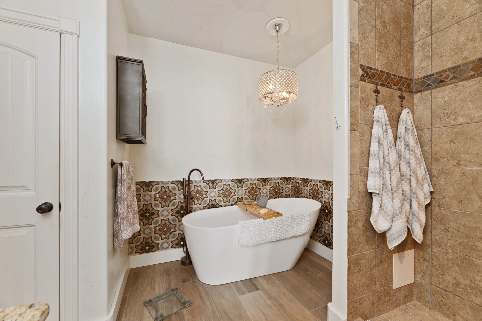 Onsuite. Freestanding soaking tub with floor-mounted bronze-finish faucet, large walkin tile accented shower, crystal chandelier, and accent wall featuring decorative tile