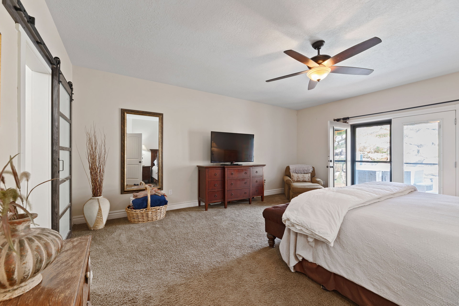 Main floor primary suite Carpeted room featuring a ceiling fan with integrated lighting, a glass-paneled barn door, and a full-length wall mirror, double doors out to back deck.