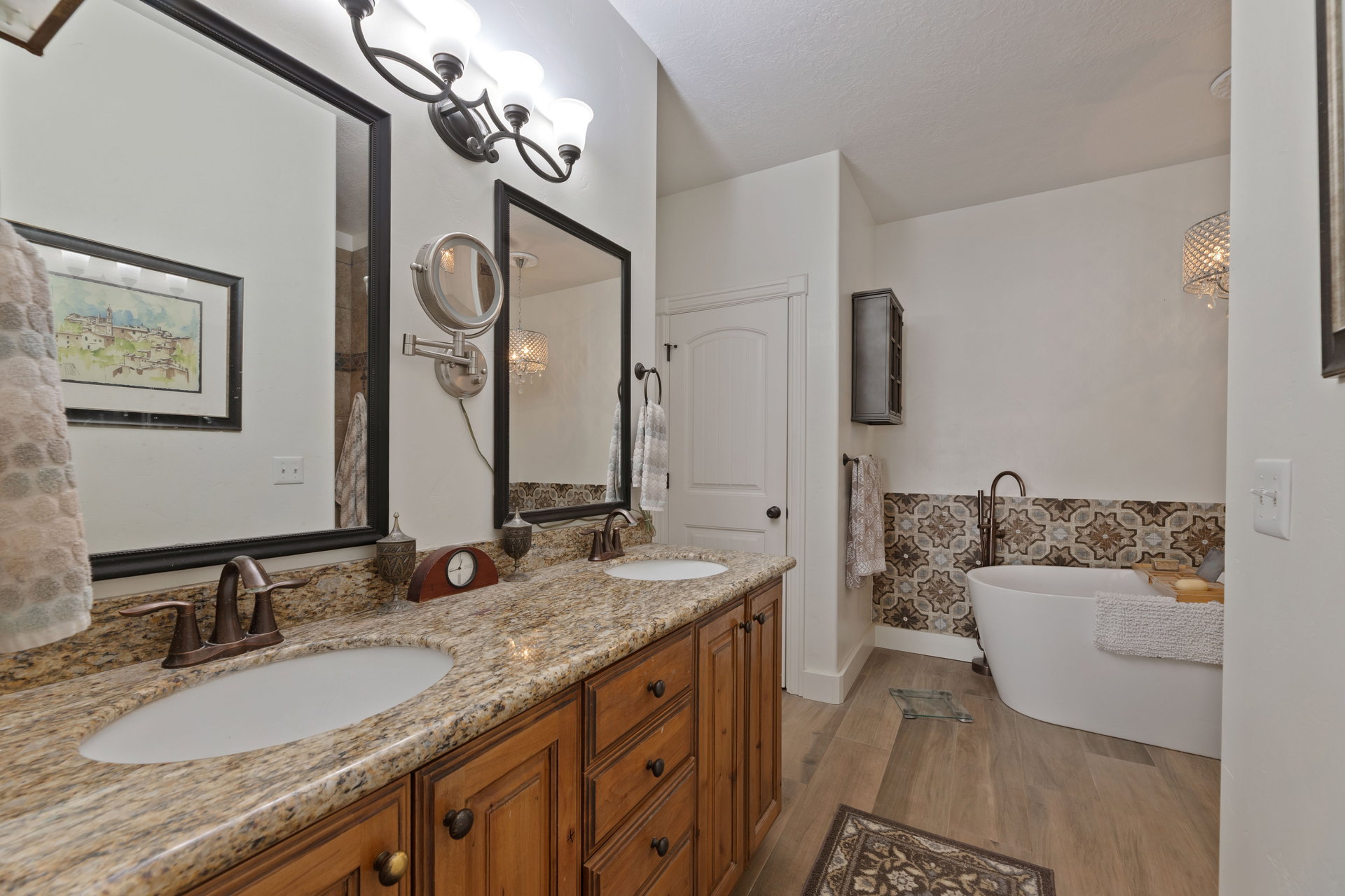 Dual vanity with granite countertop and bronze fixtures, featuring a freestanding soaking tub with patterned tile surround and a wood-finish flooring