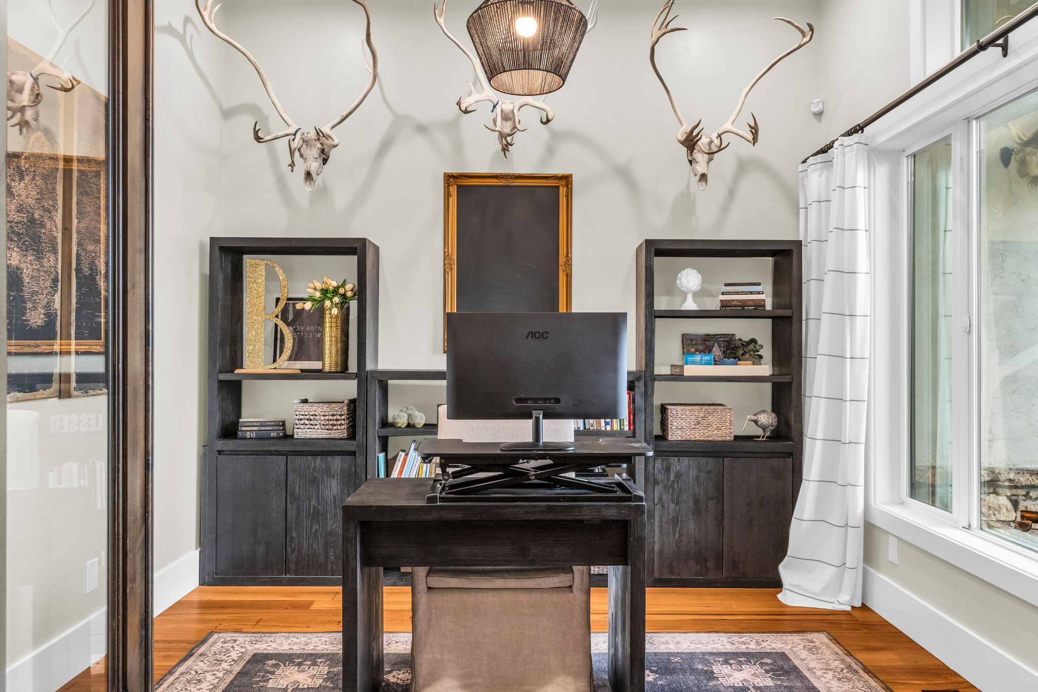 Dedicated workspace featuring wood-finish flooring, built-in shelving units with lower cabinetry, a large window with white trim, and a dark-toned suspended light fixture