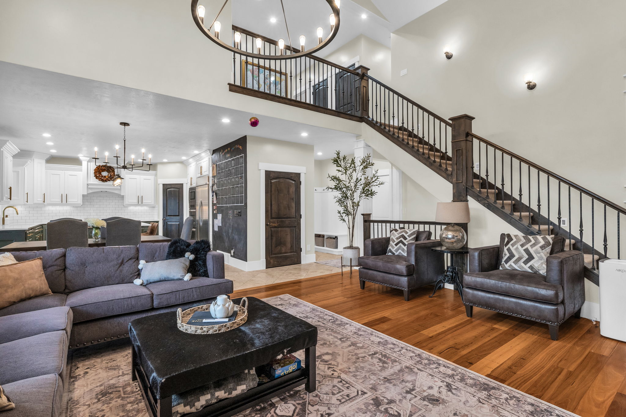 Open-concept living space featuring wood-finish flooring, a grand staircase with dark wood newel posts and metal balusters, and a contemporary chandelier