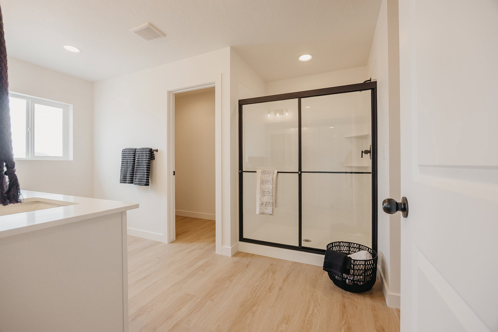 Bathroom featuring a clear glass shower enclosure with black framing, wood-finish flooring, recessed lighting, a white window, and a vanity with a light-toned countertop