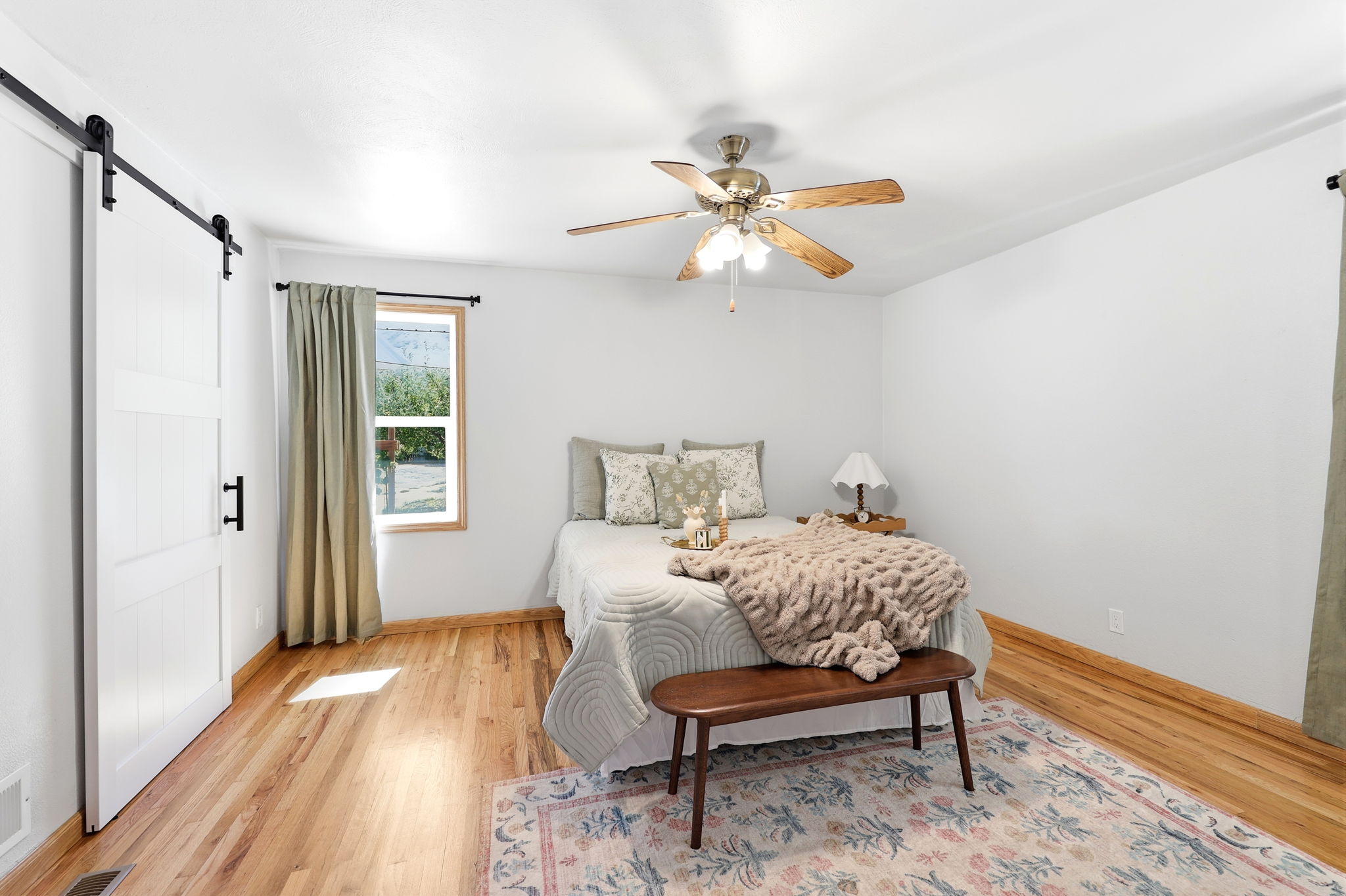 Bedroom featuring wood-finish flooring, a ceiling fan with light fixture, a window with wood trim, and a white barn door with black hardware
