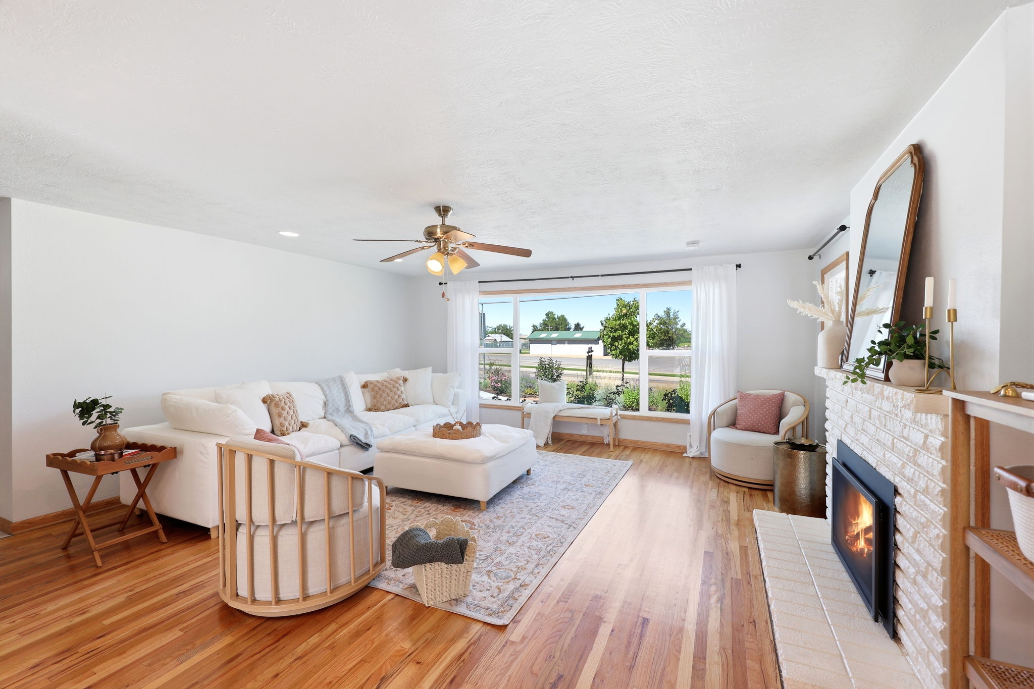 Spacious living area featuring wood-finish flooring, a white brick fireplace with a hearth, a large picture window, and a ceiling fan with integrated lighting