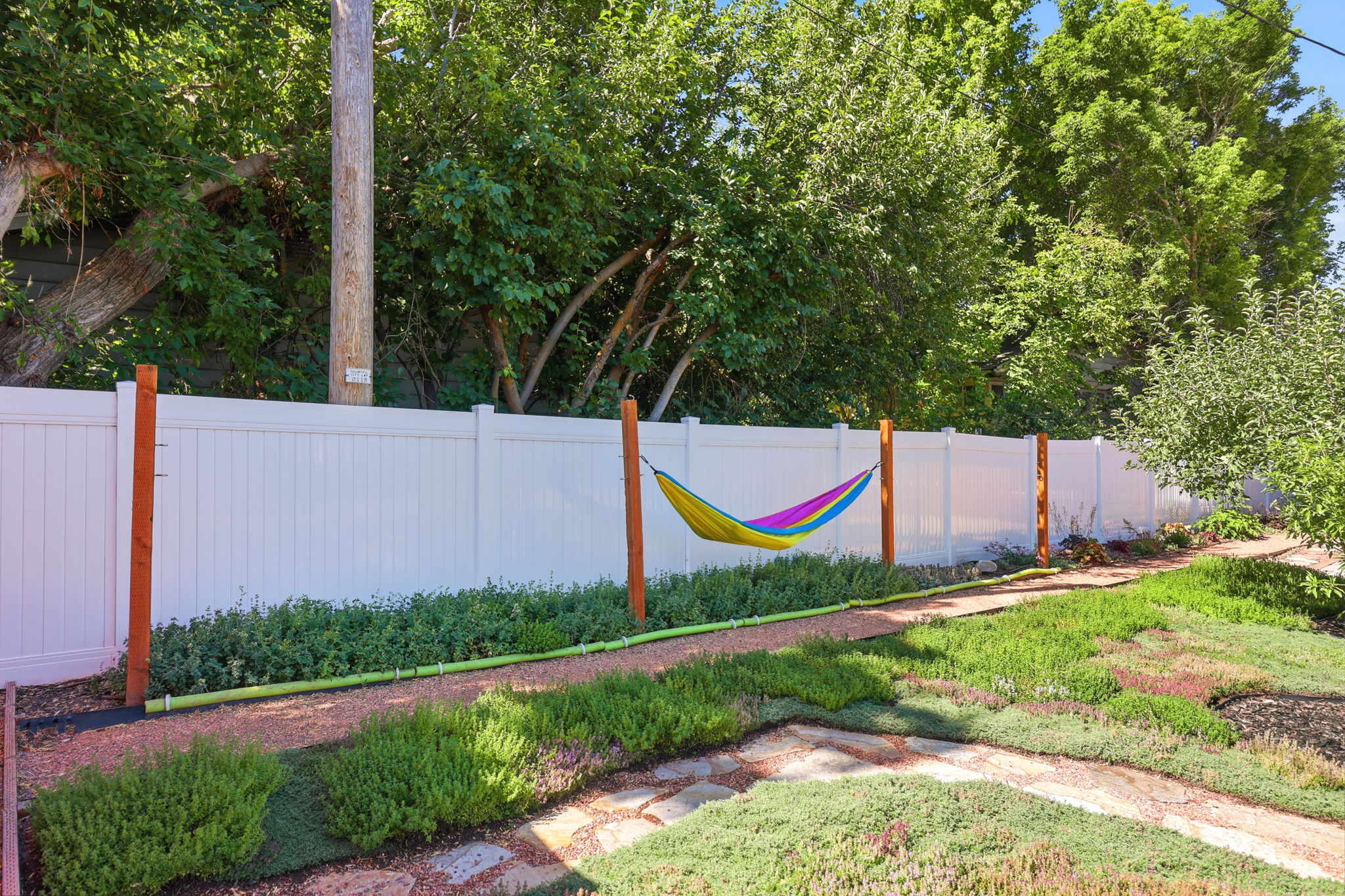 Fenced backyard featuring a gravel path, ground cover landscaping, mature trees, and a white privacy fence