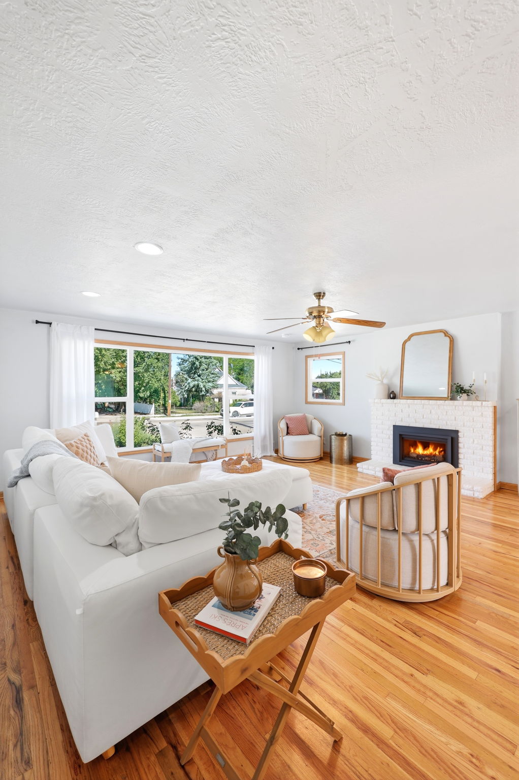 Spacious living area featuring expansive windows, wood-finish flooring, a white brick fireplace with a black insert, and a ceiling fan with integrated lighting