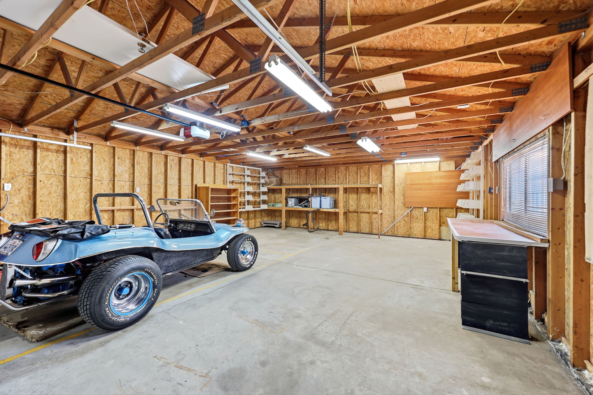 Spacious garage featuring exposed wood beam ceiling, concrete flooring, fluorescent lighting, and built-in shelving