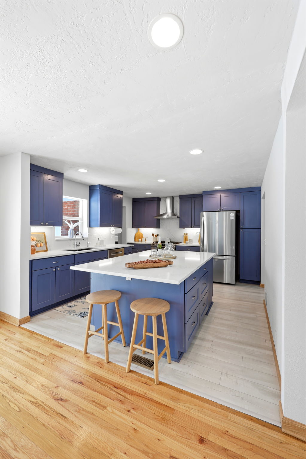 Spacious kitchen featuring a central island with white countertop, extensive blue cabinetry, stainless steel appliances, and a combination of wood and wood-finish flooring