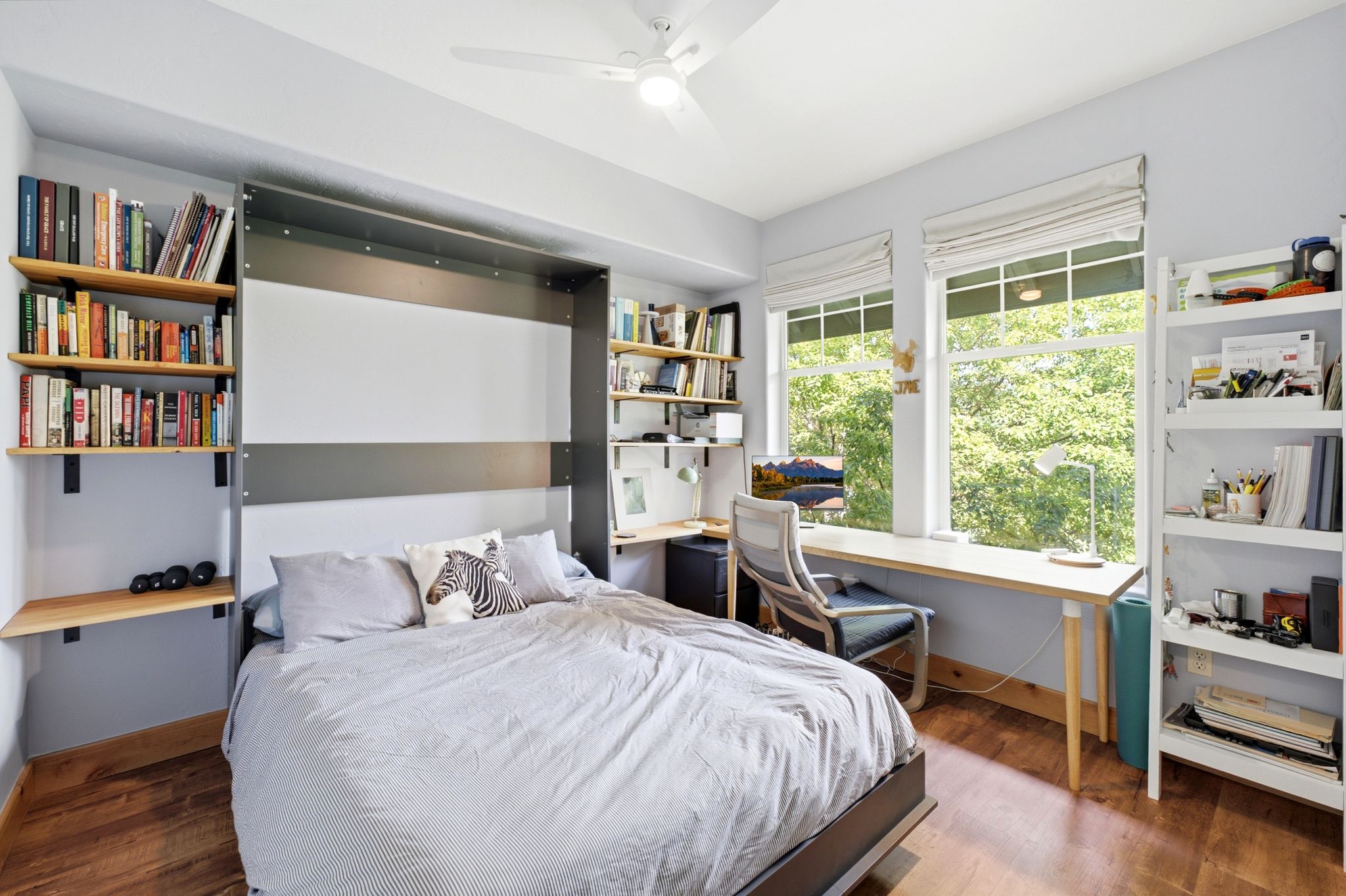 Room featuring wood-finish flooring, two large windows, a ceiling fan, built-in shelving, and a built-in desk