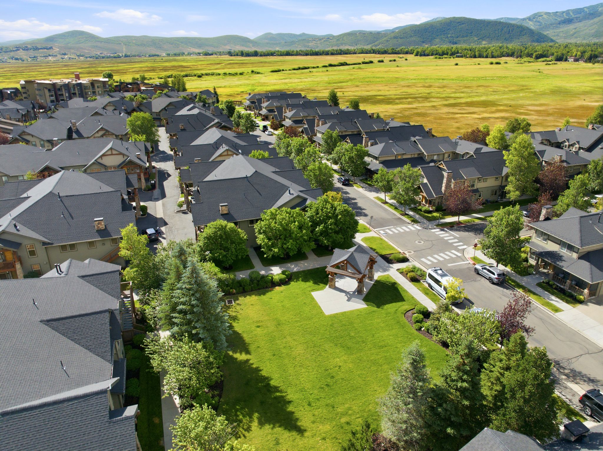 Aerial perspective showcasing a residential community with uniform dark gray shingle roofs, surrounded by lush green trees and a central green space featuring a covered pavilion