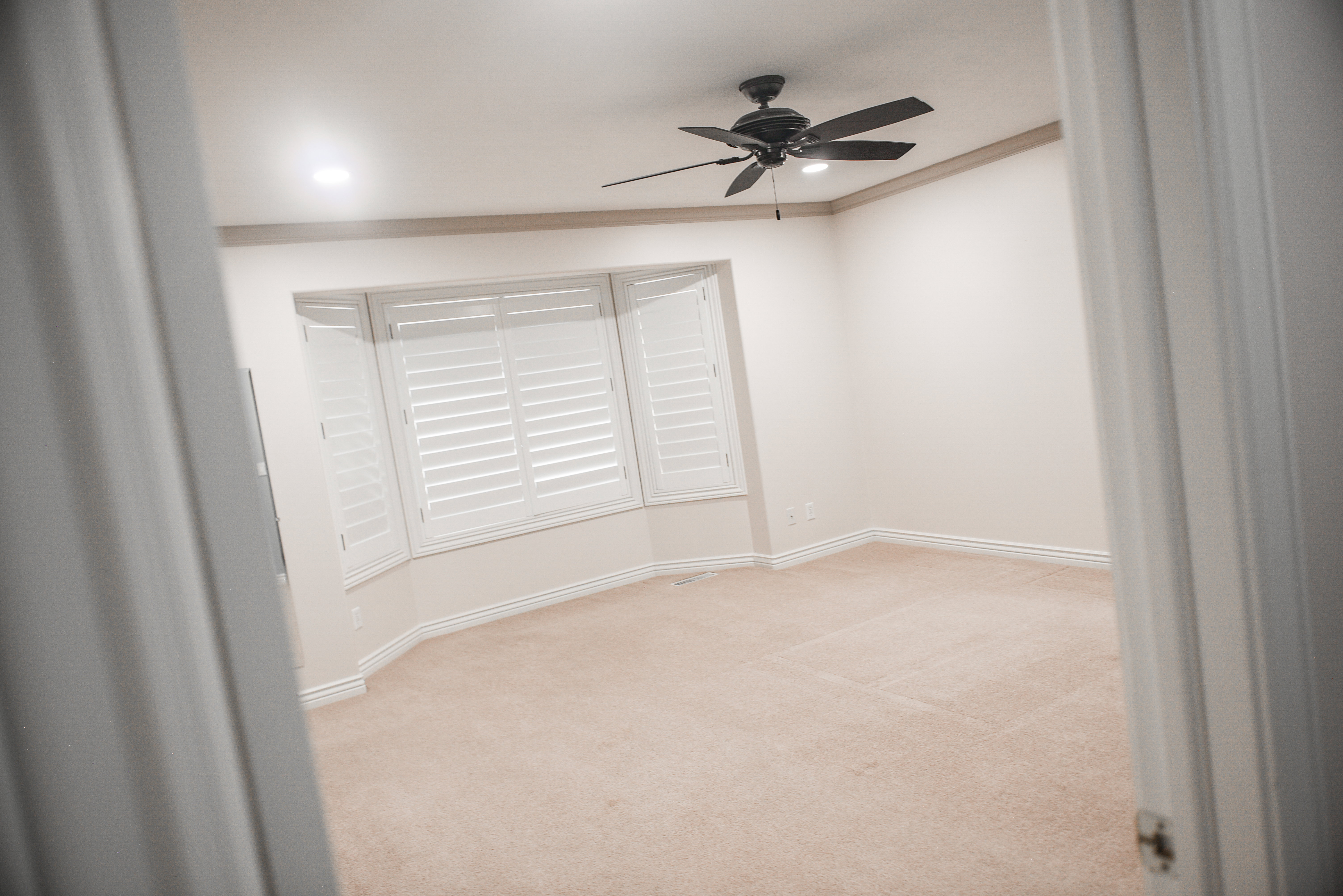 Spacious room featuring a bay window with white plantation shutters, neutral carpet, a dark ceiling fan, recessed lighting, and crown molding