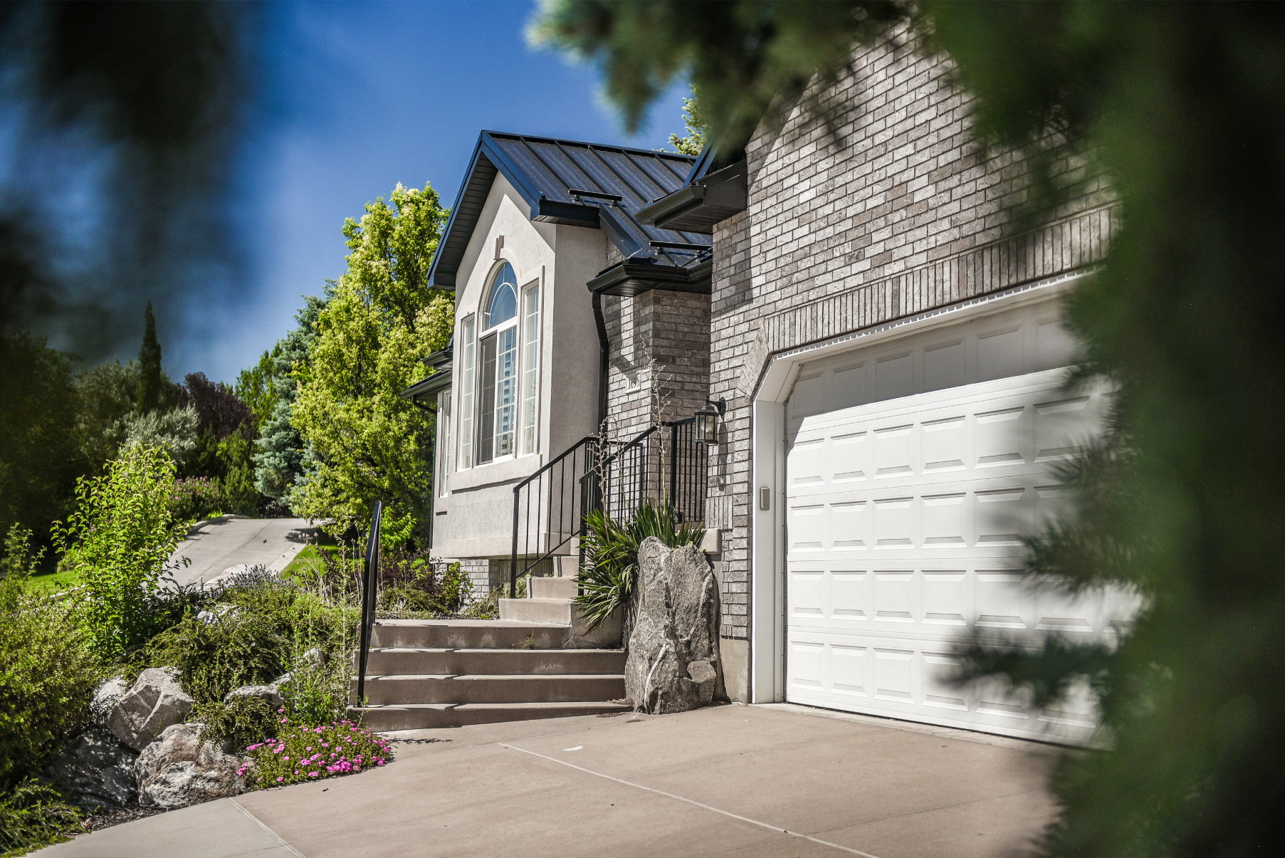 Stucco and brick exterior featuring a white paneled garage door, dark metal roof, arched window, and concrete steps with black metal railing