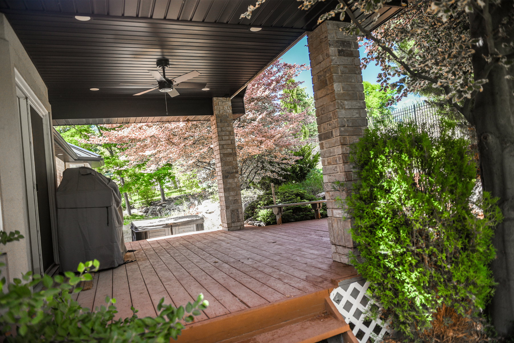 Expansive outdoor deck featuring wood-finish flooring, stone pillars, a dark wood-paneled ceiling with recessed lighting, and a ceiling fan
