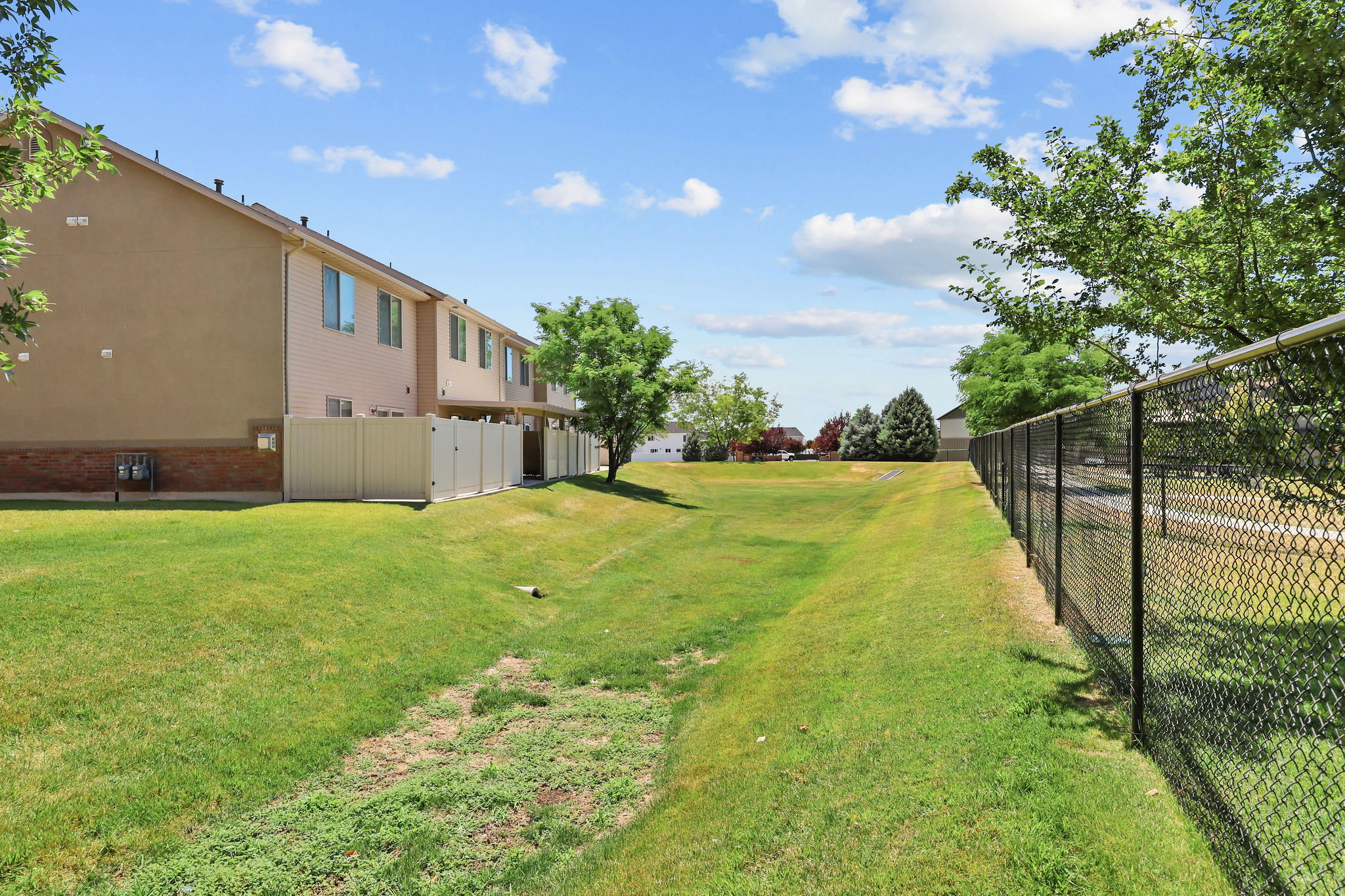 Expansive grassy area featuring a chain-link fence, mature trees, and a beige privacy fence bordering the property