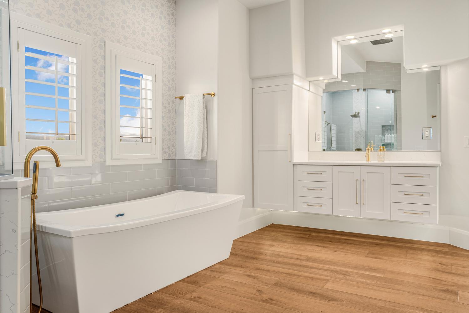 Spacious bathroom featuring a freestanding soaking tub with a floor-mounted golden faucet, dual shuttered windows, and wood-finish flooring