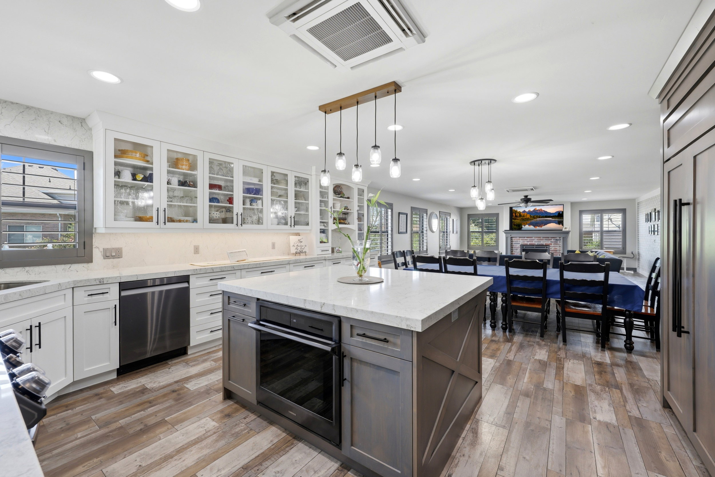 Expansive kitchen featuring a large central island with an integrated oven, wood-finish flooring, white cabinetry with glass-front uppers, stainless steel appliances, and recessed lighting