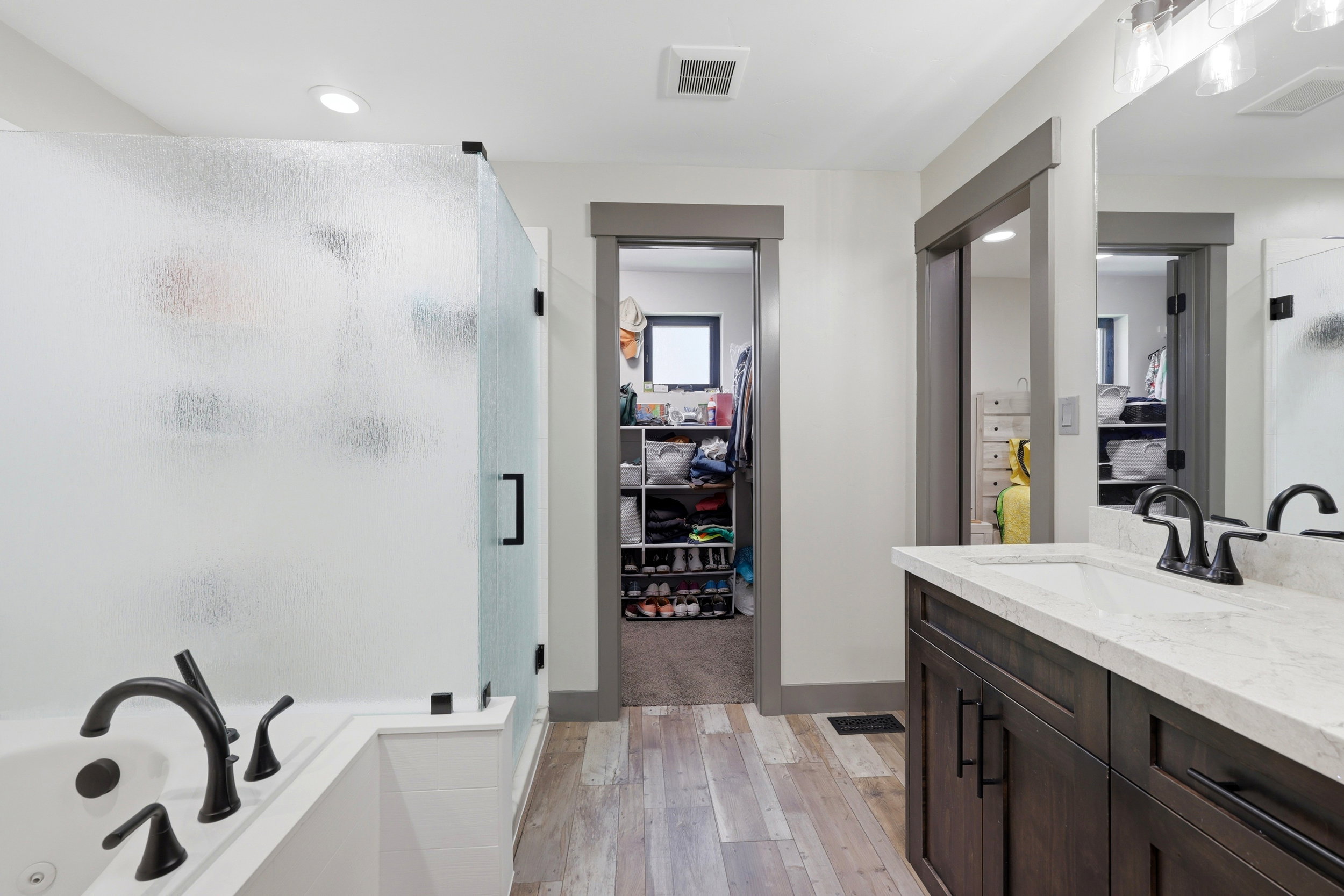 Full bathroom featuring a rectangular drop-in tub with matte black fixtures, a walk-in shower with frosted glass enclosure, a single vanity with a white countertop and undermount sink, and wood-finish flooring