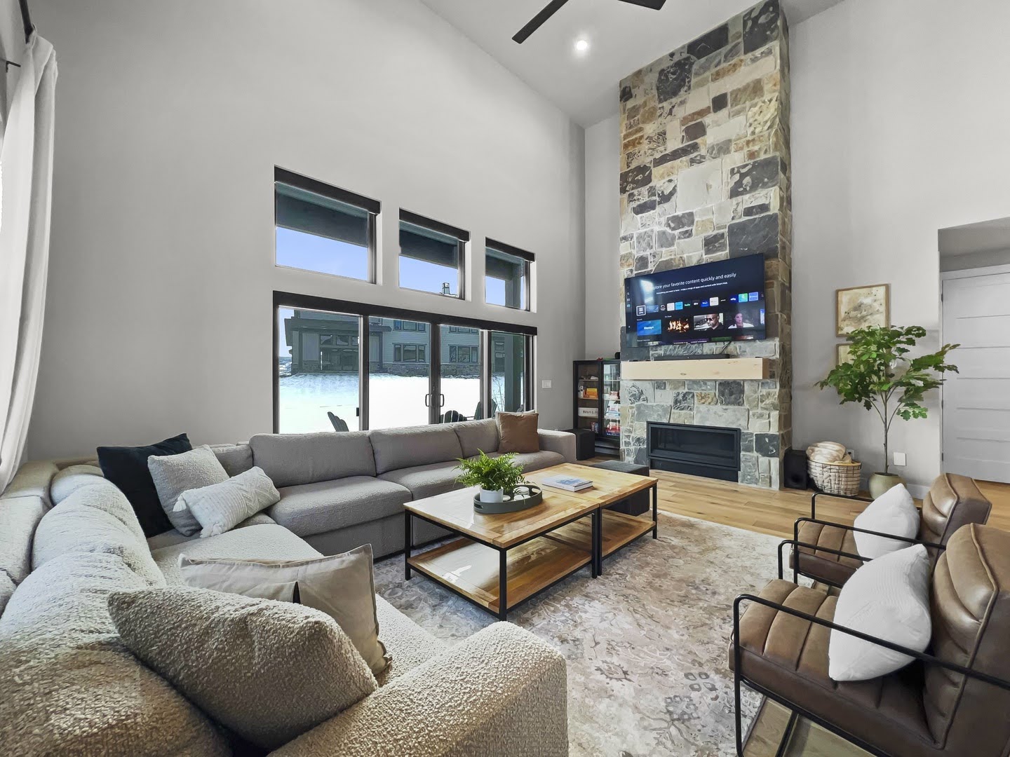 Vaulted ceiling living area featuring a floor-to-ceiling stone-faced fireplace with a wood mantle, a ceiling fan, wood-finish flooring, and expansive sliding glass doors with transom windows