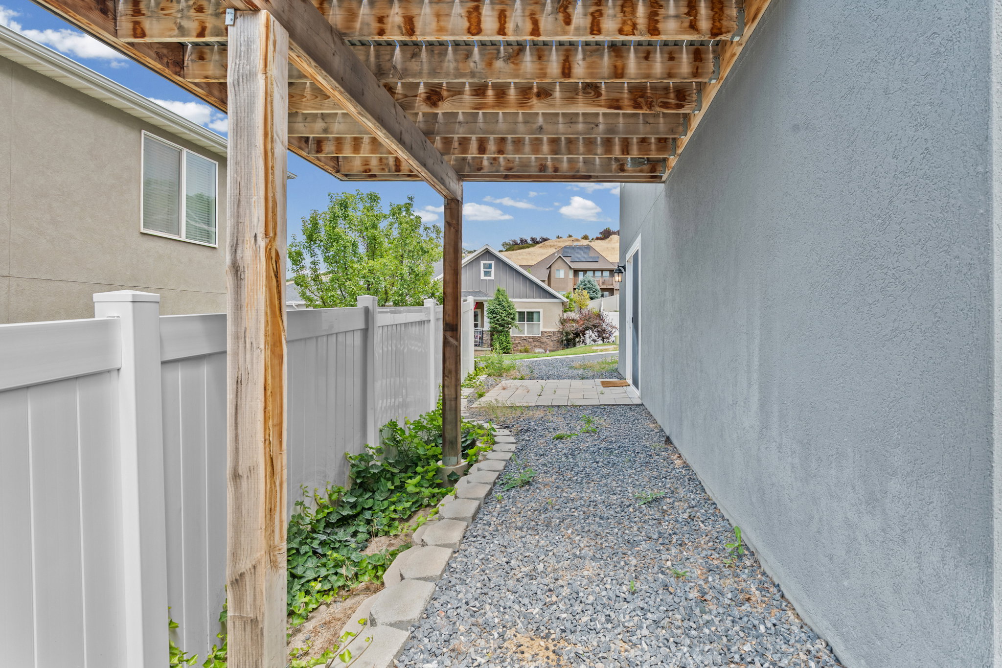 Gravel pathway under a wood-framed deck, featuring a stucco exterior wall and white privacy fencing