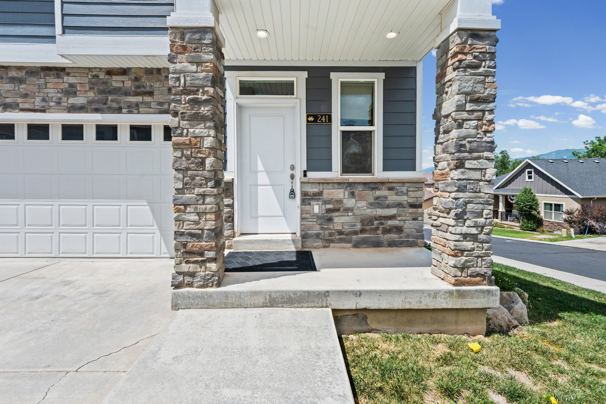 Exterior featuring a white front door with transom window, stone veneer accents, dark siding, white garage door with upper window panels, and a concrete driveway