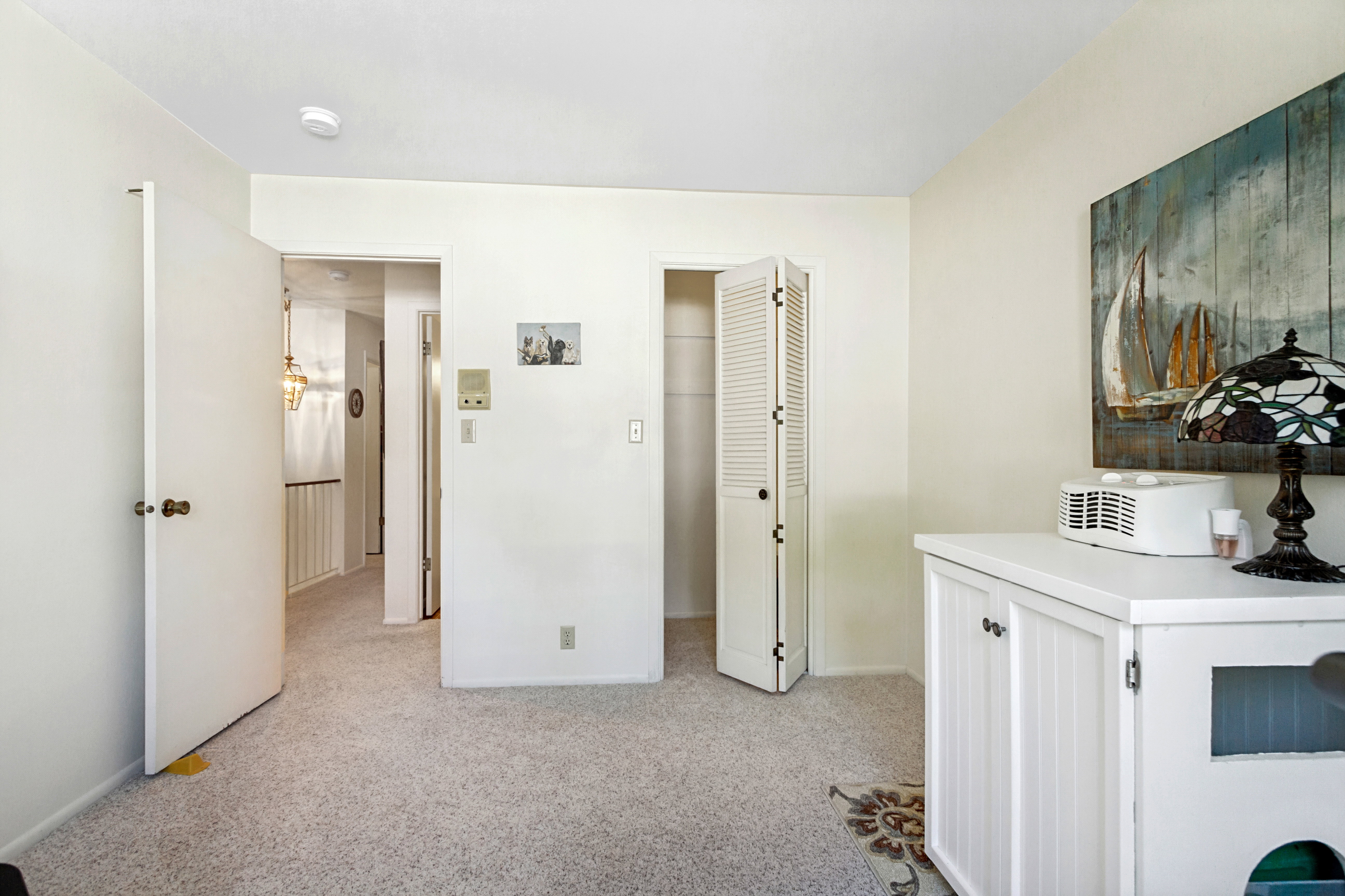 Carpeted room with white walls, featuring a louvered bi-fold closet door, white trim, and a ceiling-mounted smoke detector