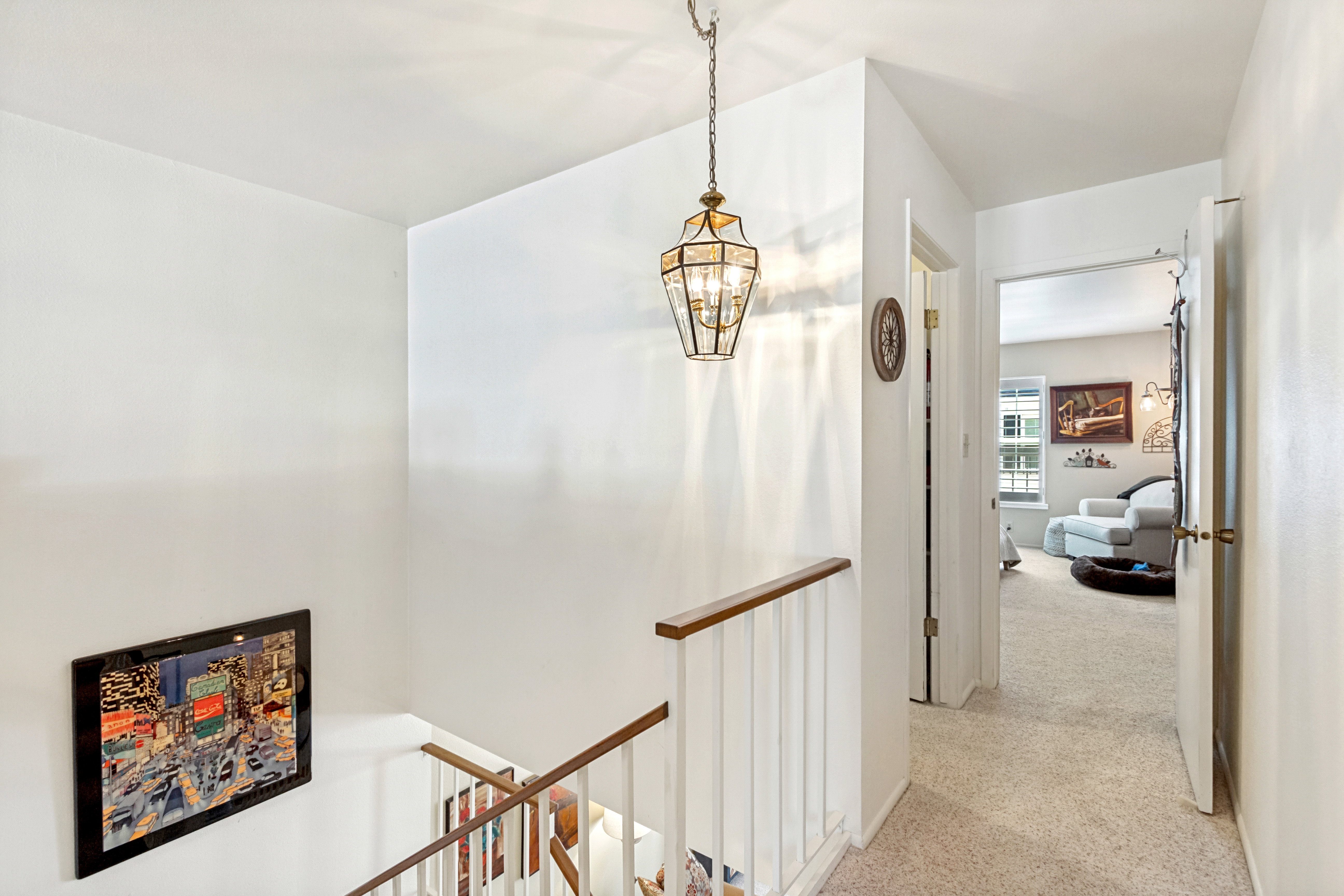 Upper-level landing featuring a hanging pendant light fixture with clear glass panels, a carpeted hallway, white walls, and a wooden handrail with white balusters