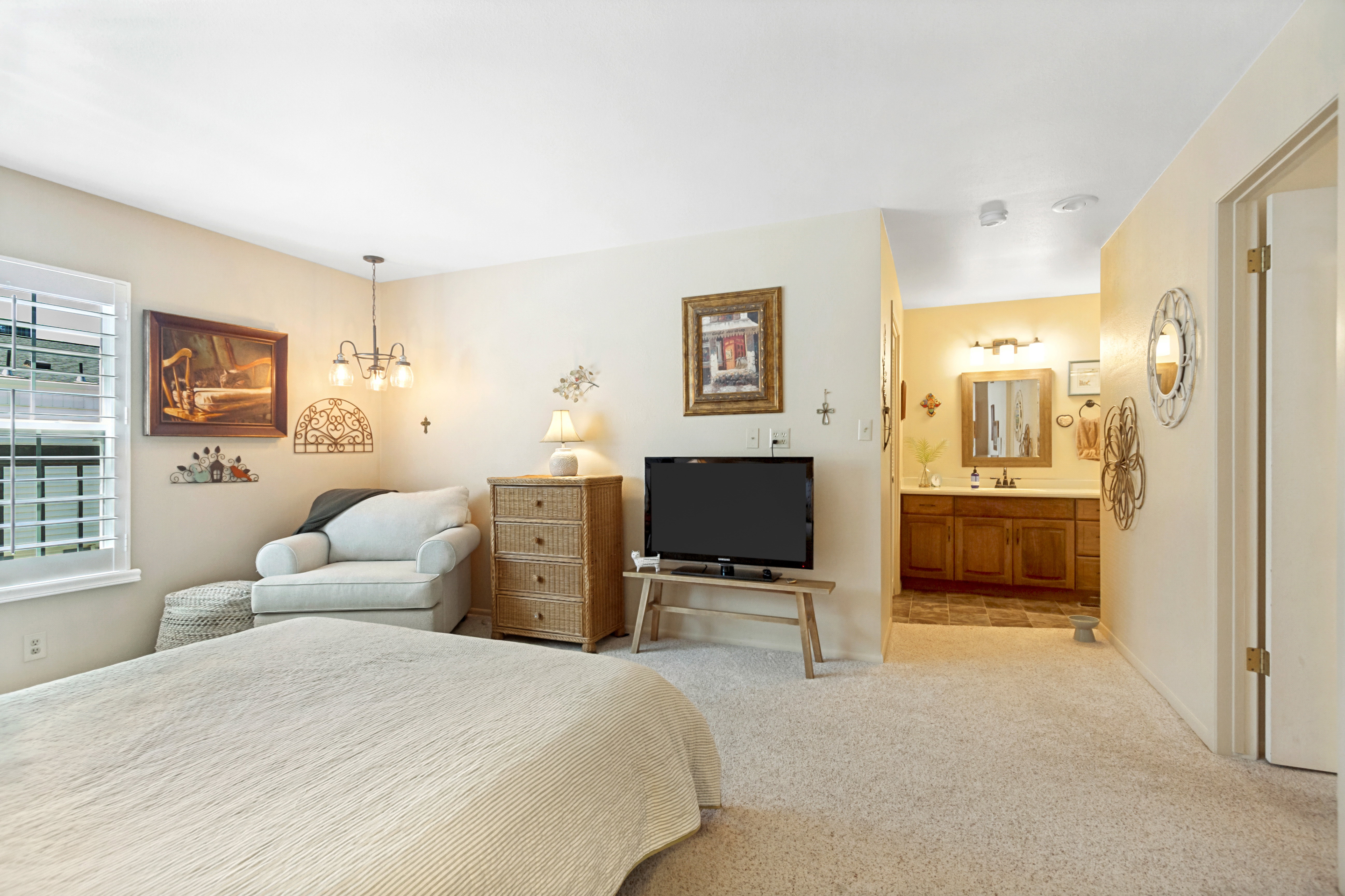 Carpeted interior space featuring a window with plantation shutters, a multi-bulb ceiling fixture, and an entryway to a bathroom with a wooden vanity