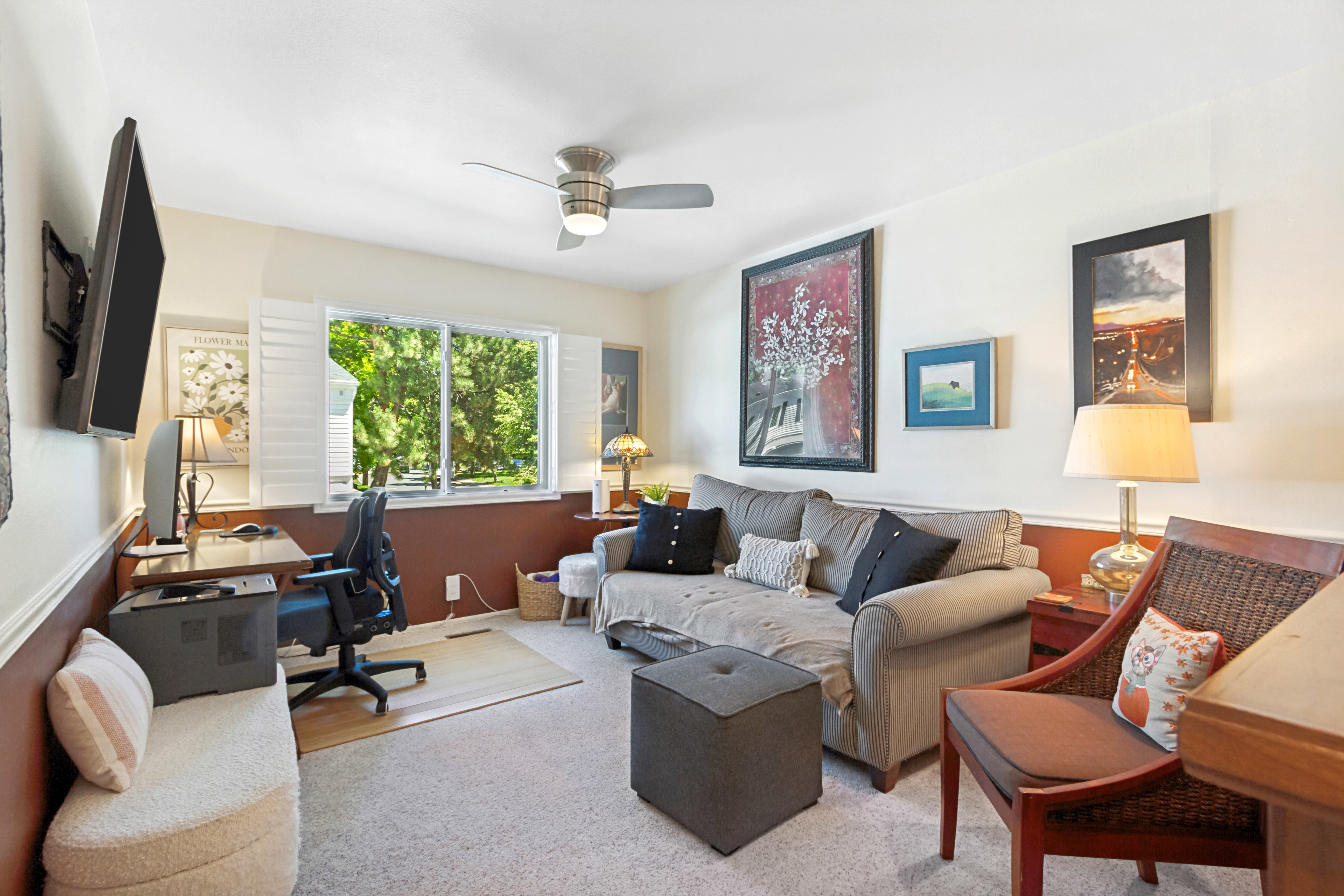 Versatile room featuring carpet flooring, natural light from a double window with white louvered shutters, a ceiling fan, and a built-in desk area